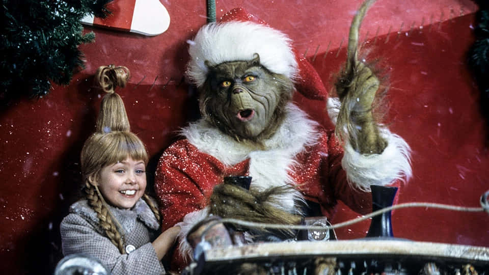 The Grinch 桌布 壁紙 — 一個小女孩在紅色雪景背景下，微笑地站在穿著聖誕老人服的鬼靈精旁邊。