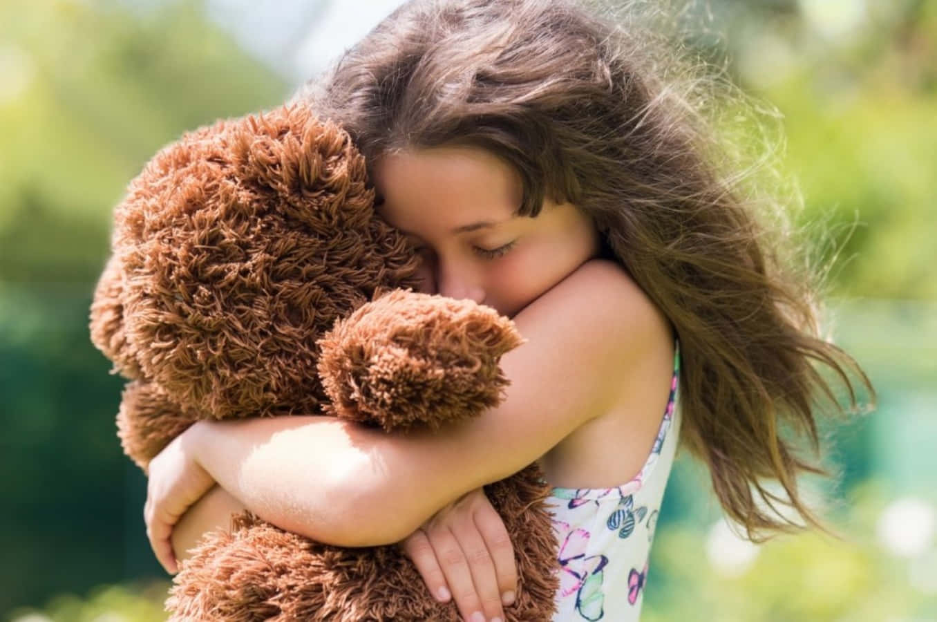 Hug 桌布 壁紙 — 一個小女孩在陽光普照的戶外環境中，緊緊擁抱一隻棕色泰迪熊，背景有模糊的綠色植物。