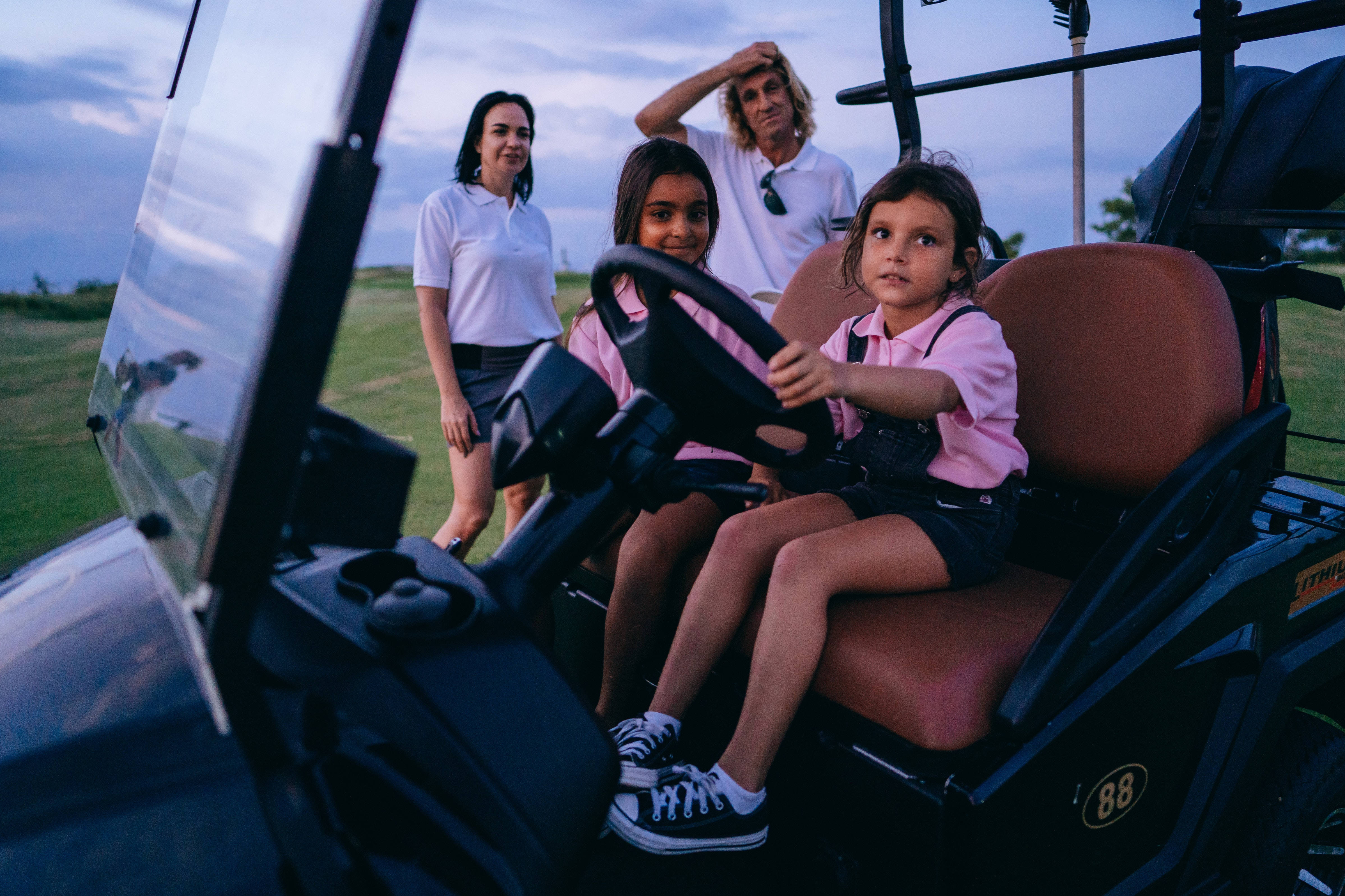 4K Golf 桌布 壁紙 — 一個小女孩抓住高爾夫球車的方向盤，她的家人在黃昏的高爾夫球場上從後面觀看。
