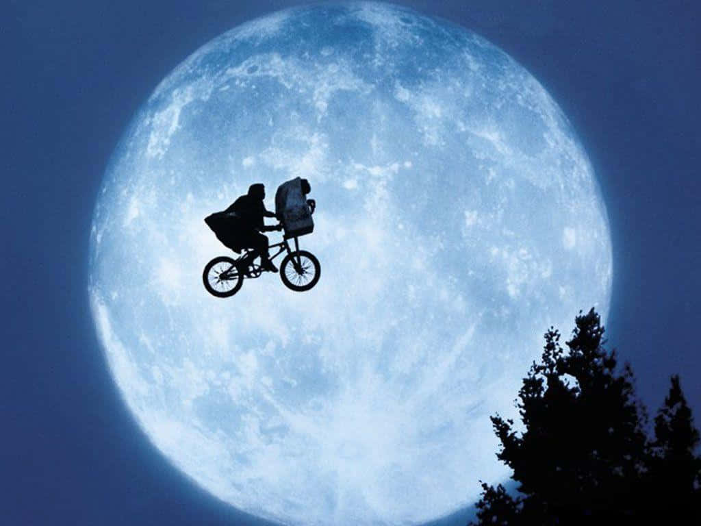 Et 桌布 壁紙 — 一個小孩和一隻狗以剪影形式騎腳踏車，背景是一個巨大的發光藍色月亮，下方可見樹梢。