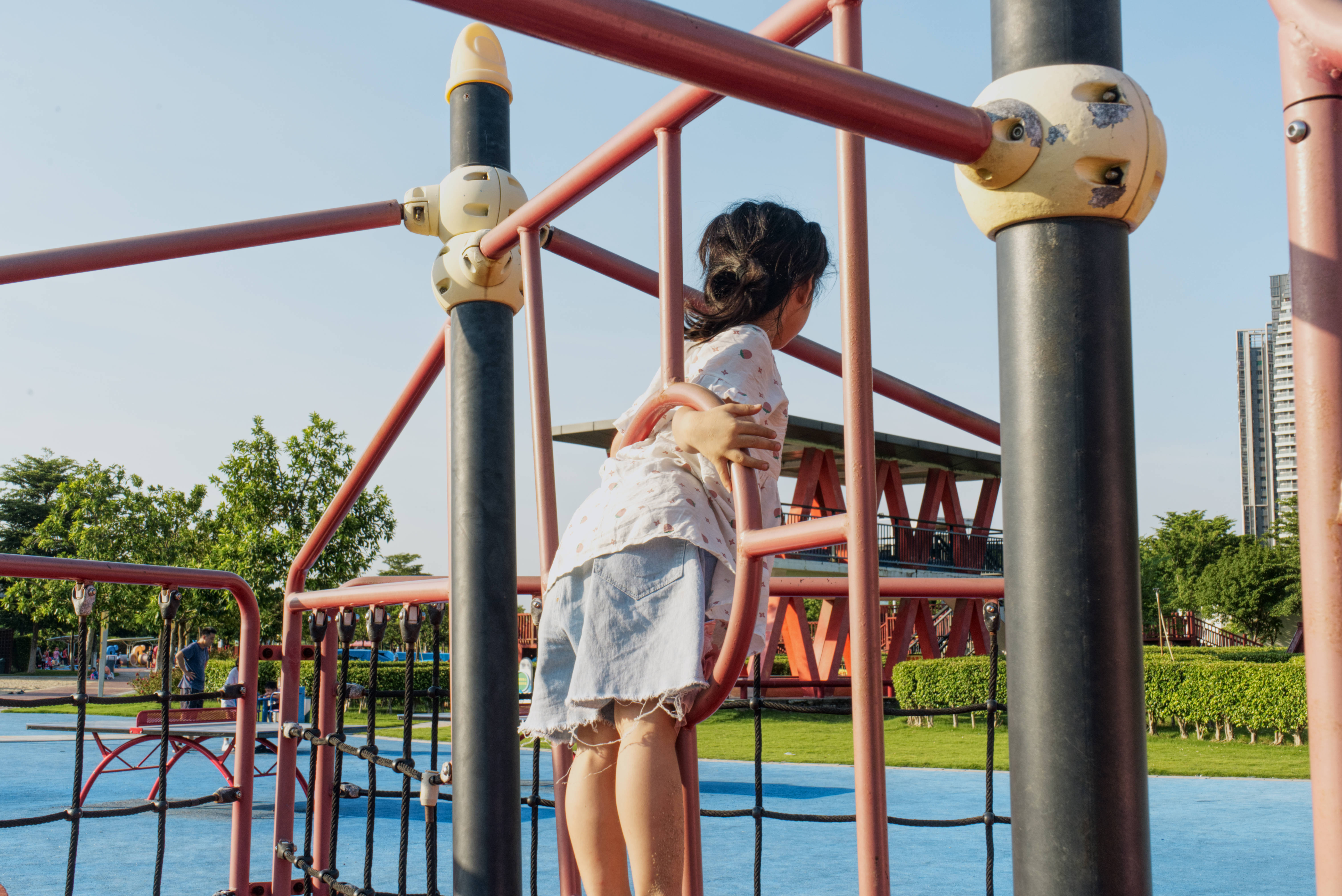 Playground 桌布 壁紙 — 一個小孩在明亮的日光下攀爬紅色遊樂場結構，背景有綠樹和城市天際線。