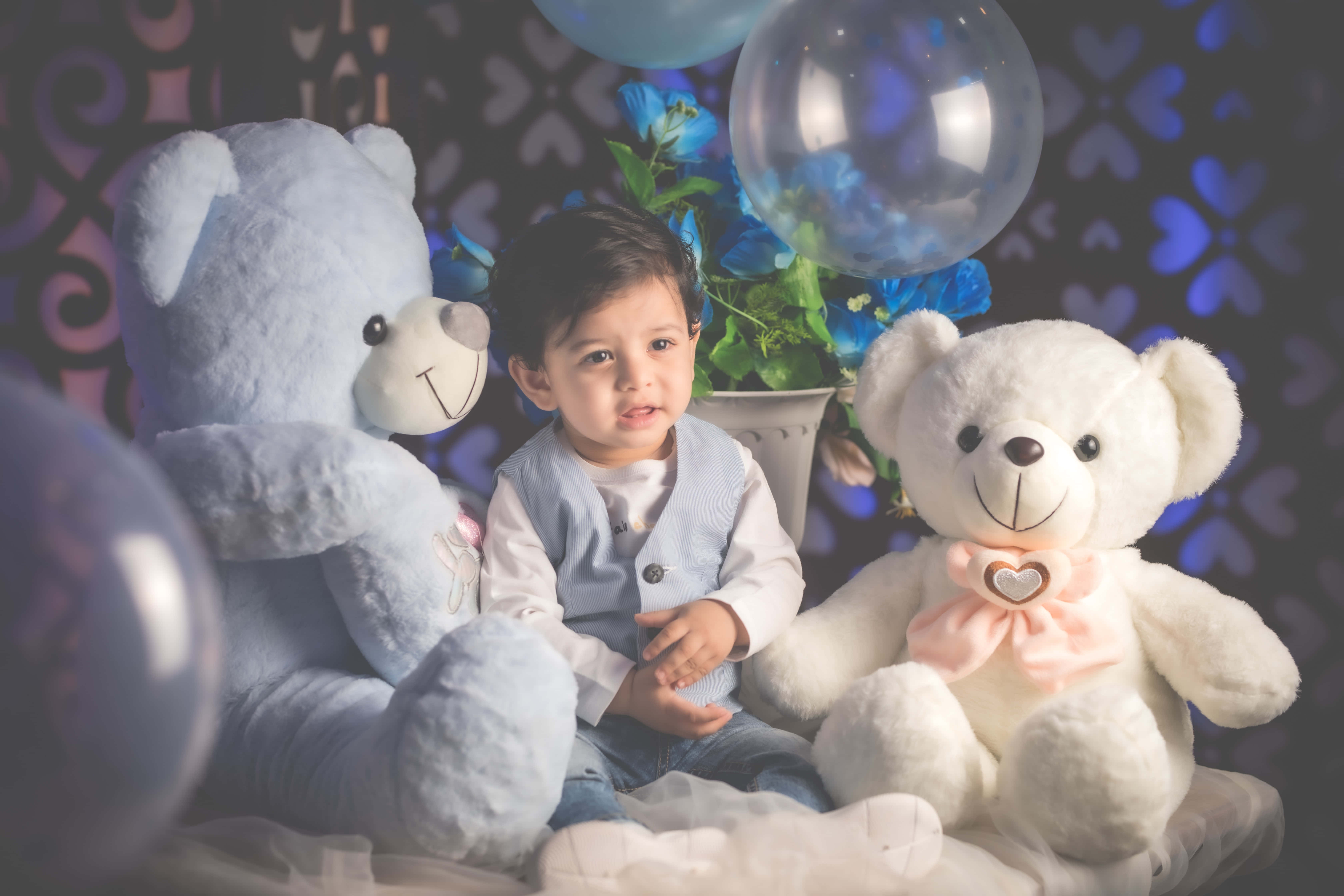 Baby Boy 桌布 壁紙 — 一個小孩坐在兩隻大泰迪熊之間，周圍環繞著藍色氣球和花卉裝飾，在柔和照明的攝影棚環境中。