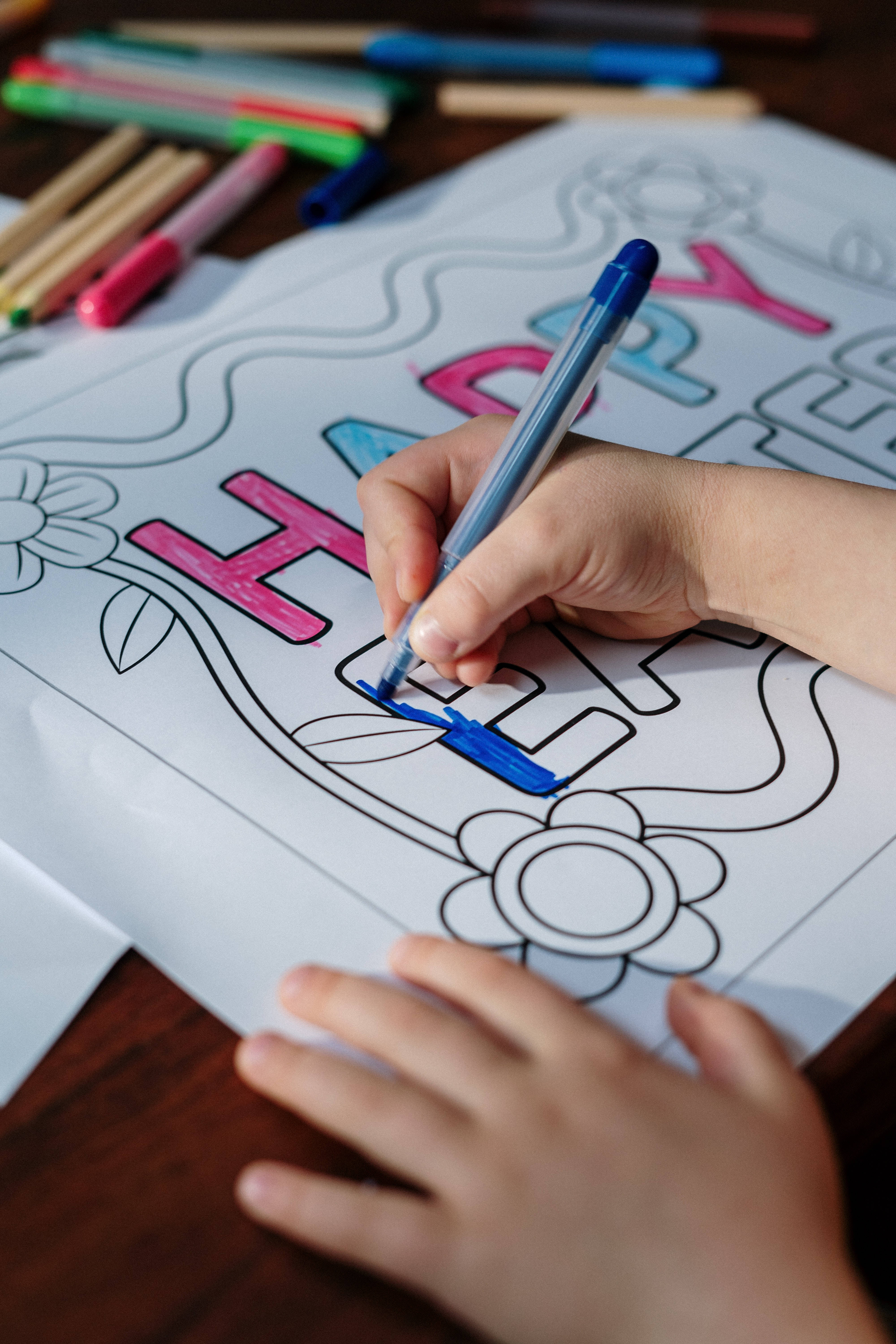 復活節 桌布 壁紙 — 一個小孩的手正在用藍色馬克筆為「HAPPY」著色頁中的字母H上色，周圍散落著蠟筆和馬克筆。