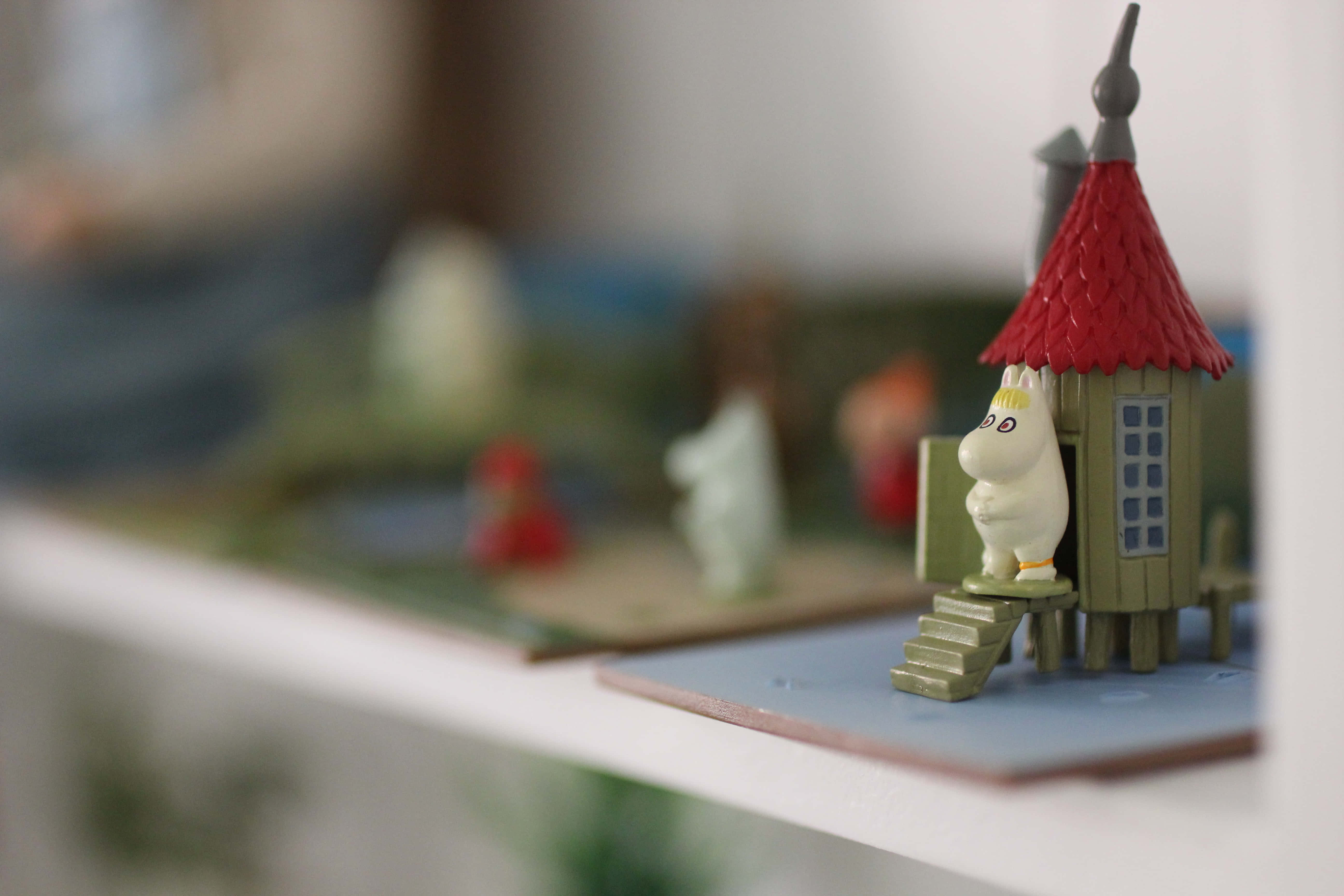 Moomin Desktop 桌布 壁紙 — 一個小小的姆明公仔站在架上一個紅屋頂綠屋前的階梯上，背景柔和模糊。