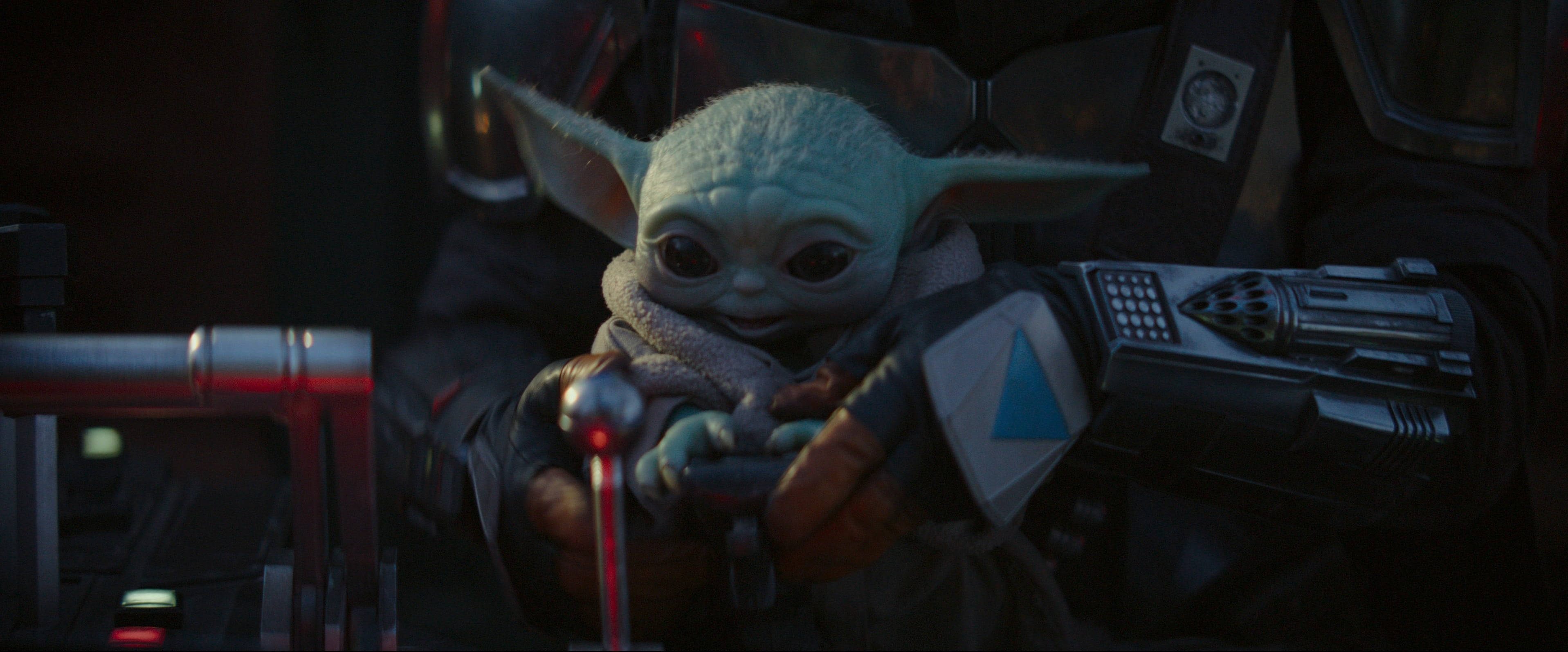 Mandalorian And Baby Yoda 桌布 壁紙 — 一個小小的綠色Baby Yoda緊握著一個發光的紅色光劍柄，同時依偎在一個黑暗裝甲人物的懷抱中。