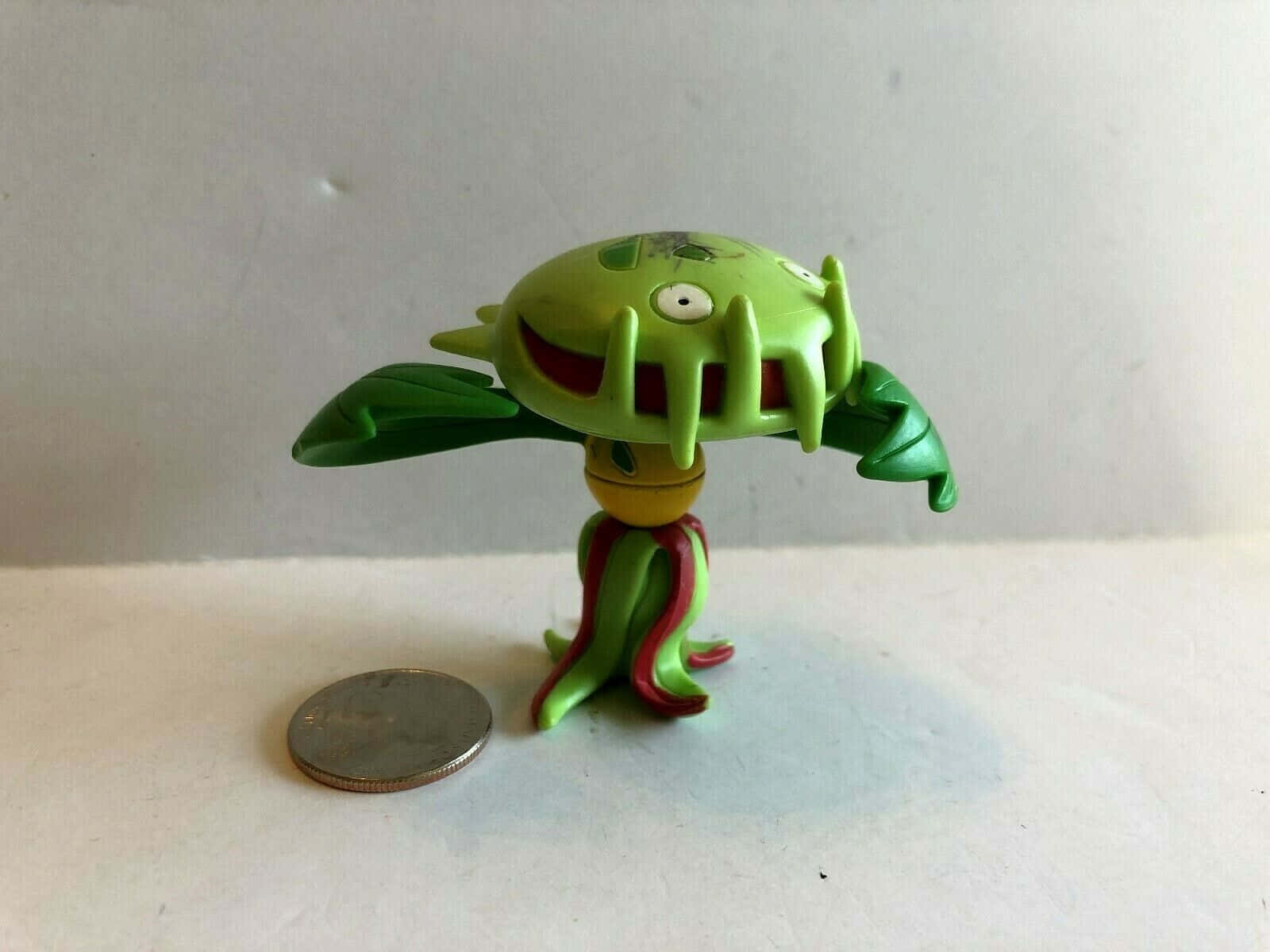 3D 桌布 壁紙 — 一個小小的綠色塑膠公仔，造型是卡通植物怪獸，有樹葉手臂，旁邊有一枚硬幣作為比例參考。
