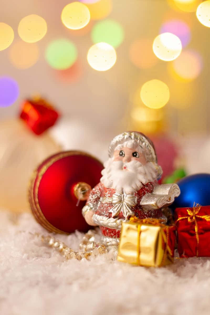 Santa 桌布 壁紙 — 一個小小的聖誕老人公仔坐在節日裝飾品和禮物之間，放在雪白的絨毛上，背後有彩色散景燈光柔和地發光。