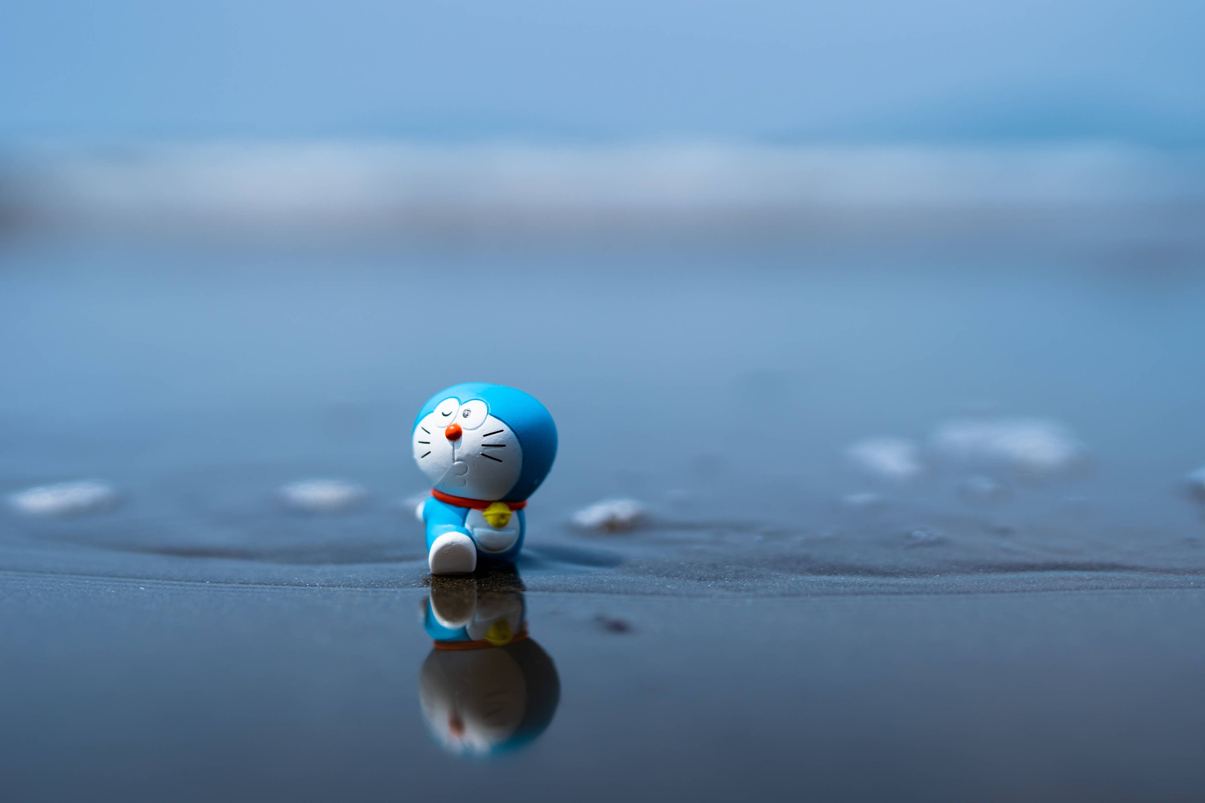Doraemon 桌布 壁紙 — 一個小小的藍色哆啦A夢公仔獨自坐在濕沙上，背景是模糊的海洋，並有清晰的倒影。