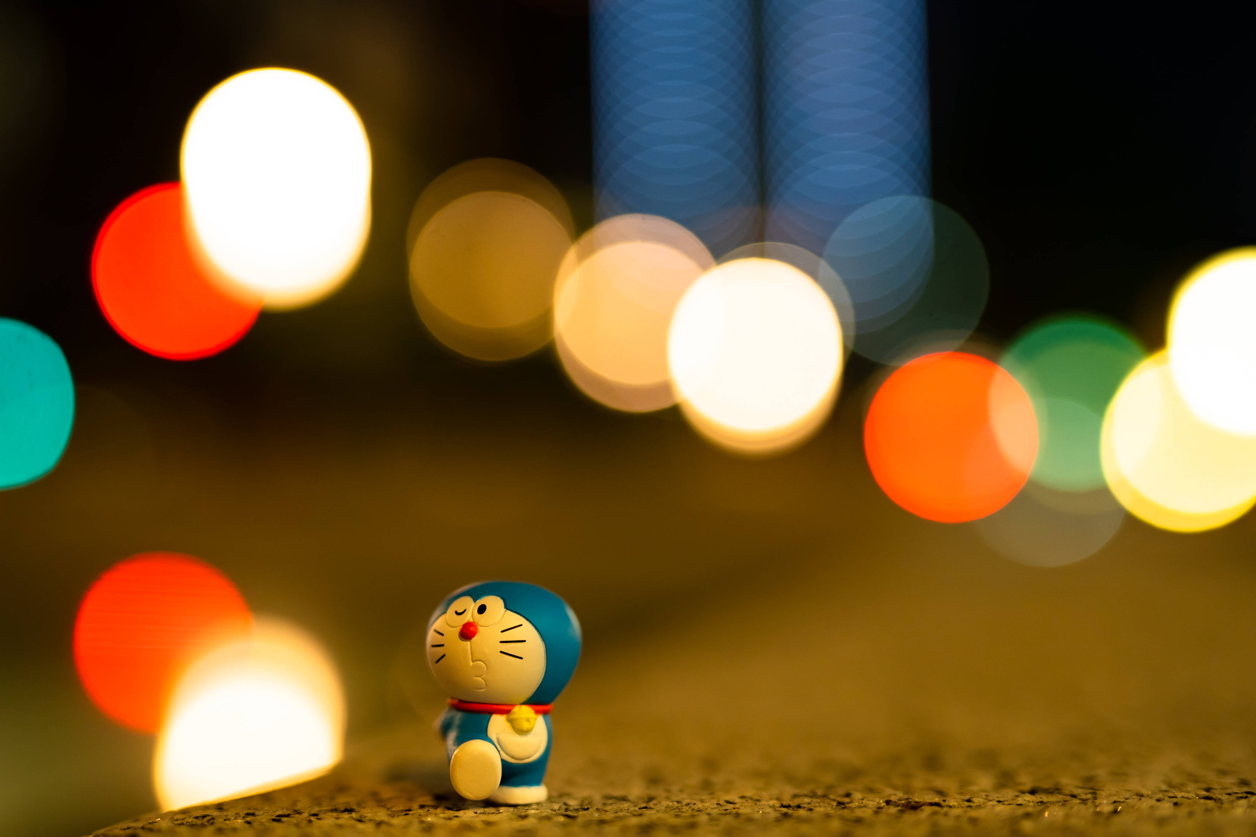 Doraemon 桌布 壁紙 — 一個小小的藍色哆啦A夢公仔獨自坐在有紋理的表面上，背景是充滿活力的散景夜景城市景觀。