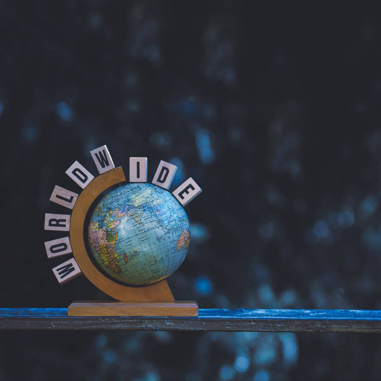 World Globe 桌布 壁紙 — 一個小木製地球儀，上面有 'WORLDWIDE' 字母積木拱形排列，放在一個深色架子上，背景是模糊的藍色散景。