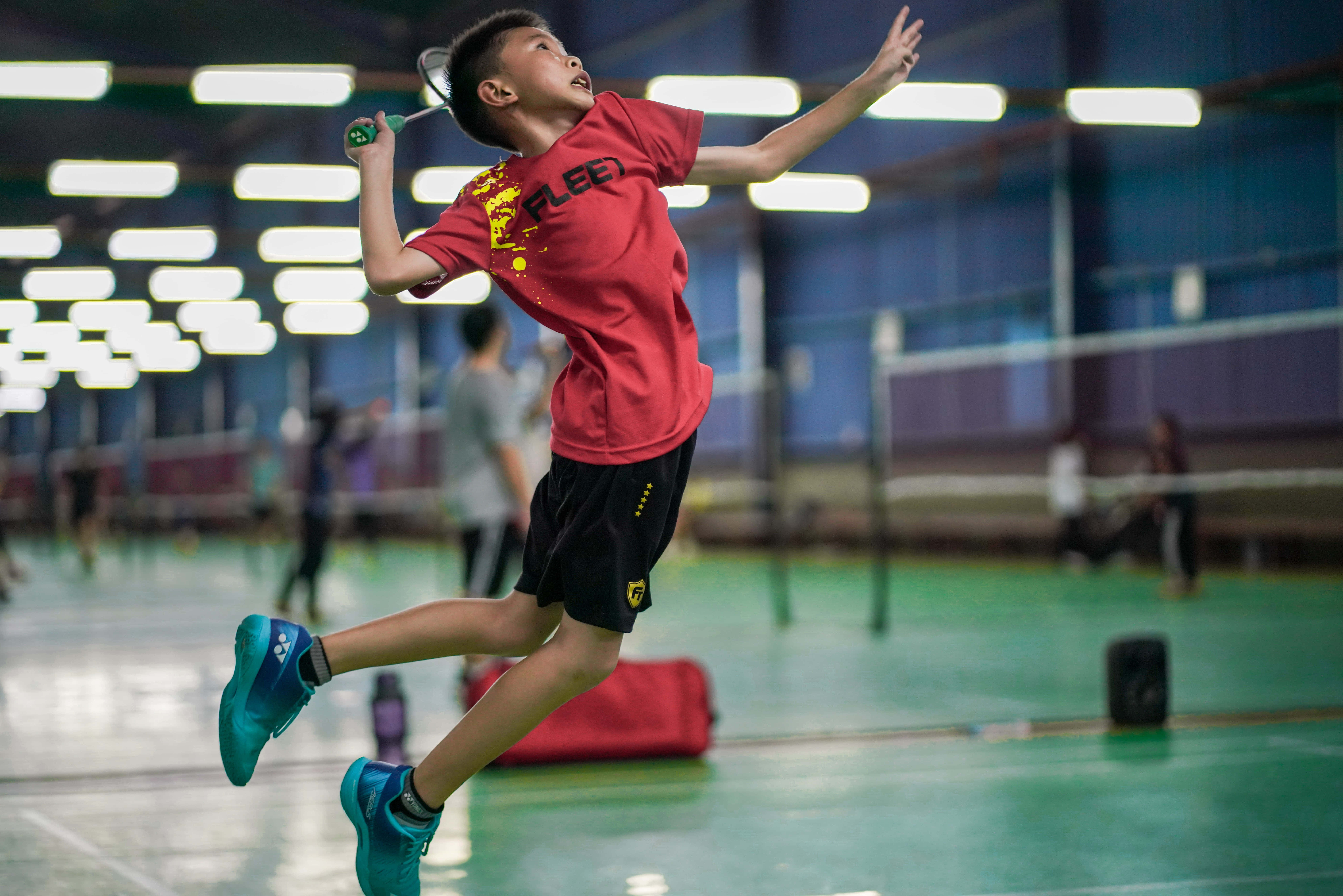 Badminton 桌布 壁紙 — 一個小男孩在光線充足的室內球場中，動態地躍起揮動羽毛球拍。