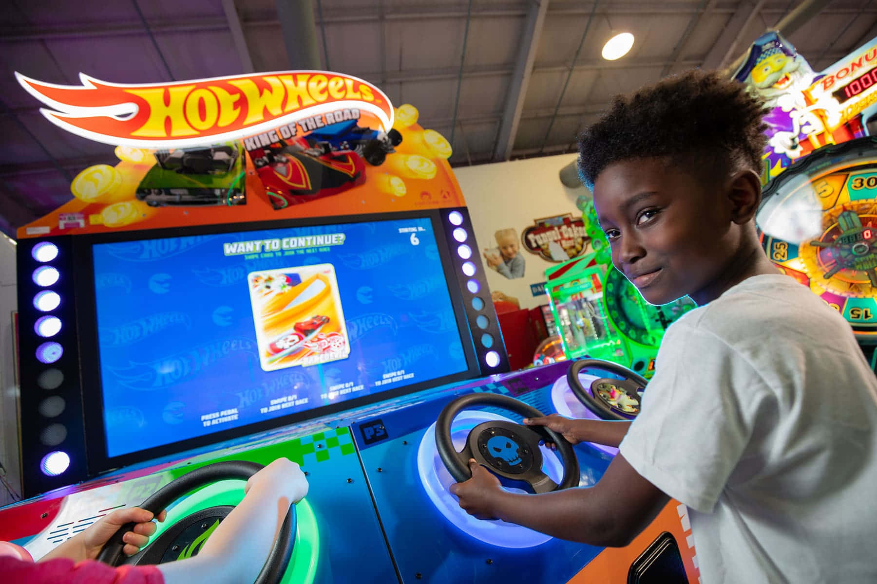 Arcade Games 桌布 壁紙 — 一個小男孩在光線明亮的遊戲機中心玩Hot Wheels賽車遊戲時微笑。
