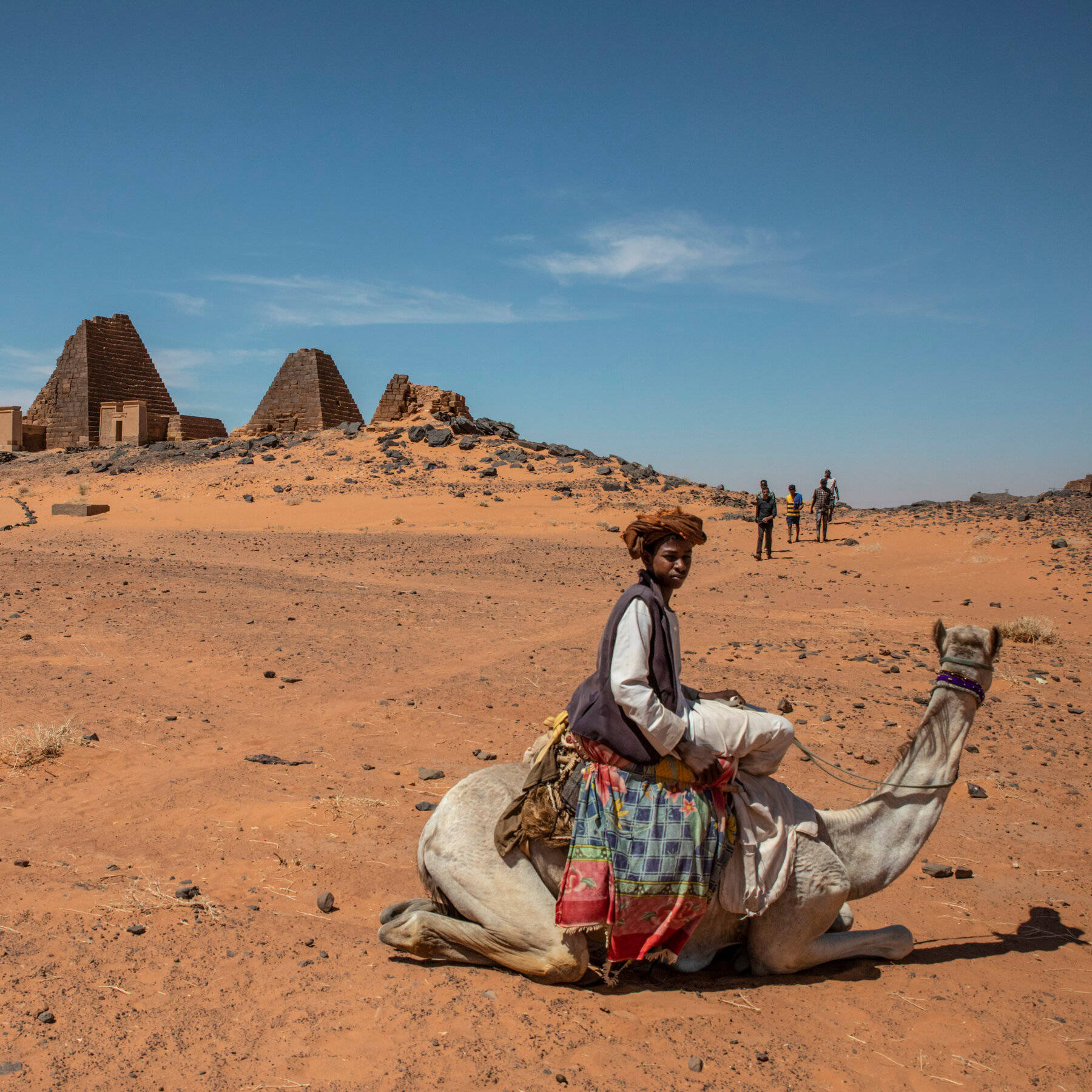 Sudan 桌布 壁紙 — 一個小男孩坐在一隻休息的駱駝上，身處廣闊的沙漠，背景有古老的金字塔在清澈的藍天下聳立。