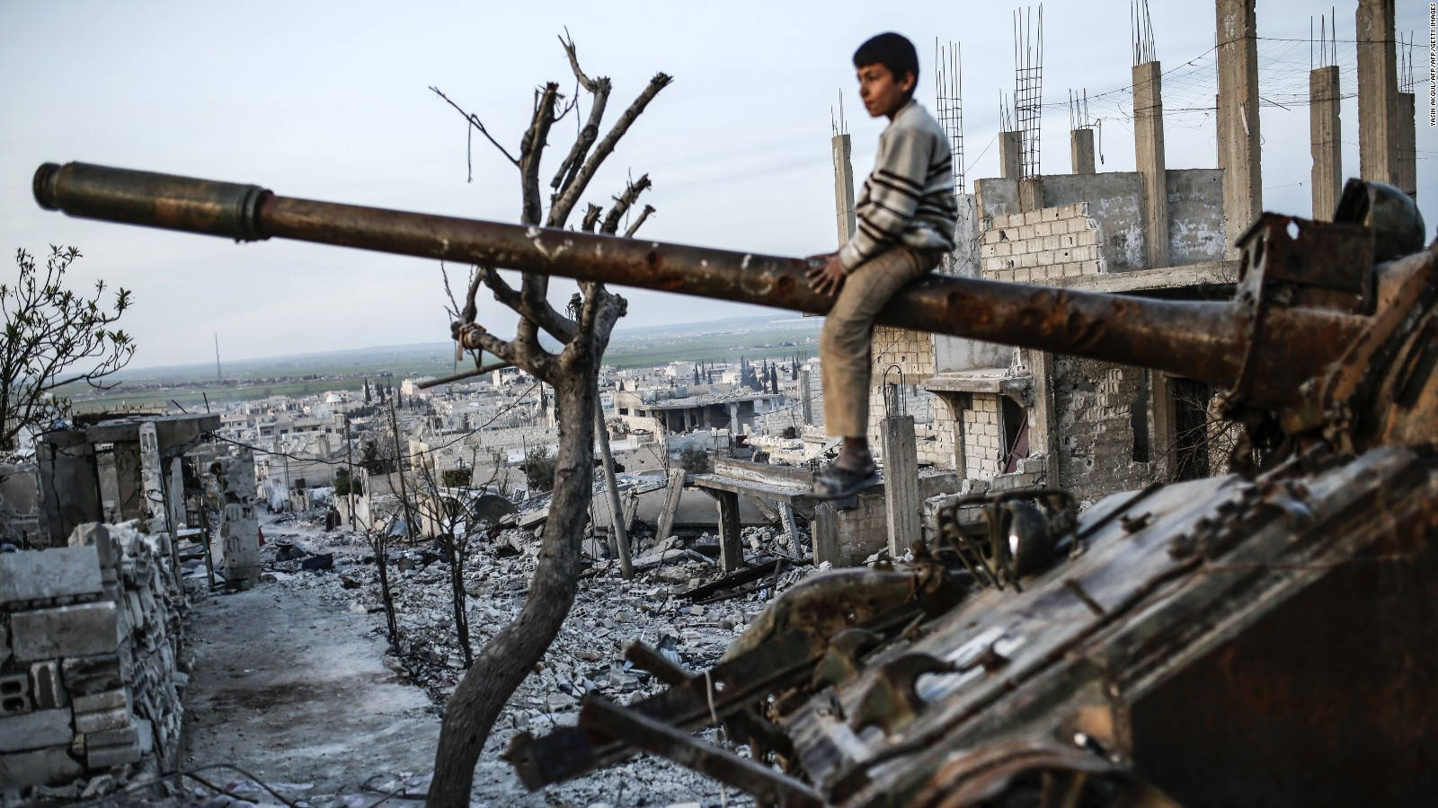 Syria 桌布 壁紙 — 一個小男孩坐在生鏽的坦克炮管上，身處一個被戰爭摧毀的城市景觀中，到處是瓦礫和只剩下骨架的建築物。