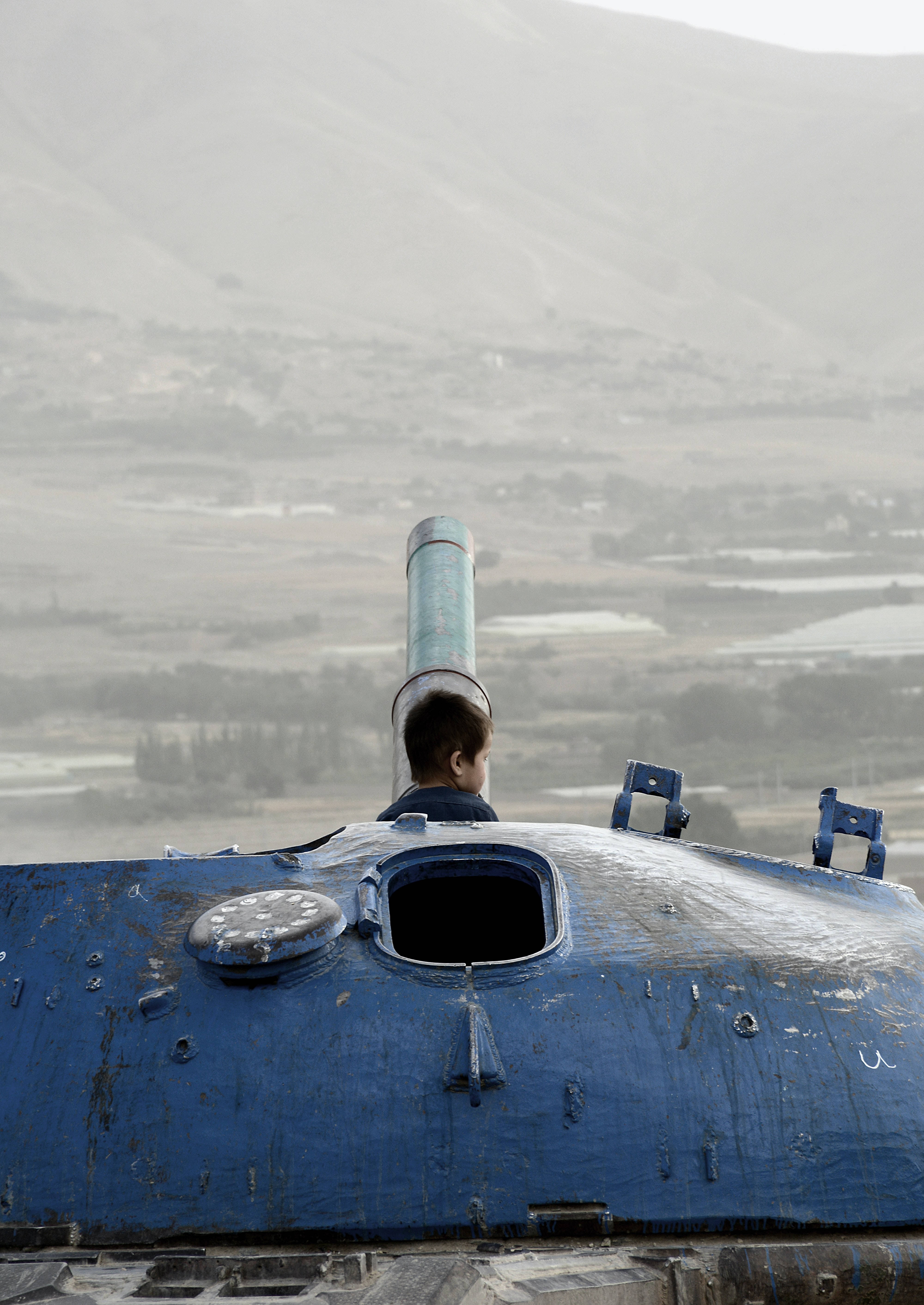 Kabul 桌布 壁紙 — 一個小男孩從一個飽經風霜的藍色坦克艙口探出頭來，俯瞰著煙霧瀰漫的山谷和遠山。