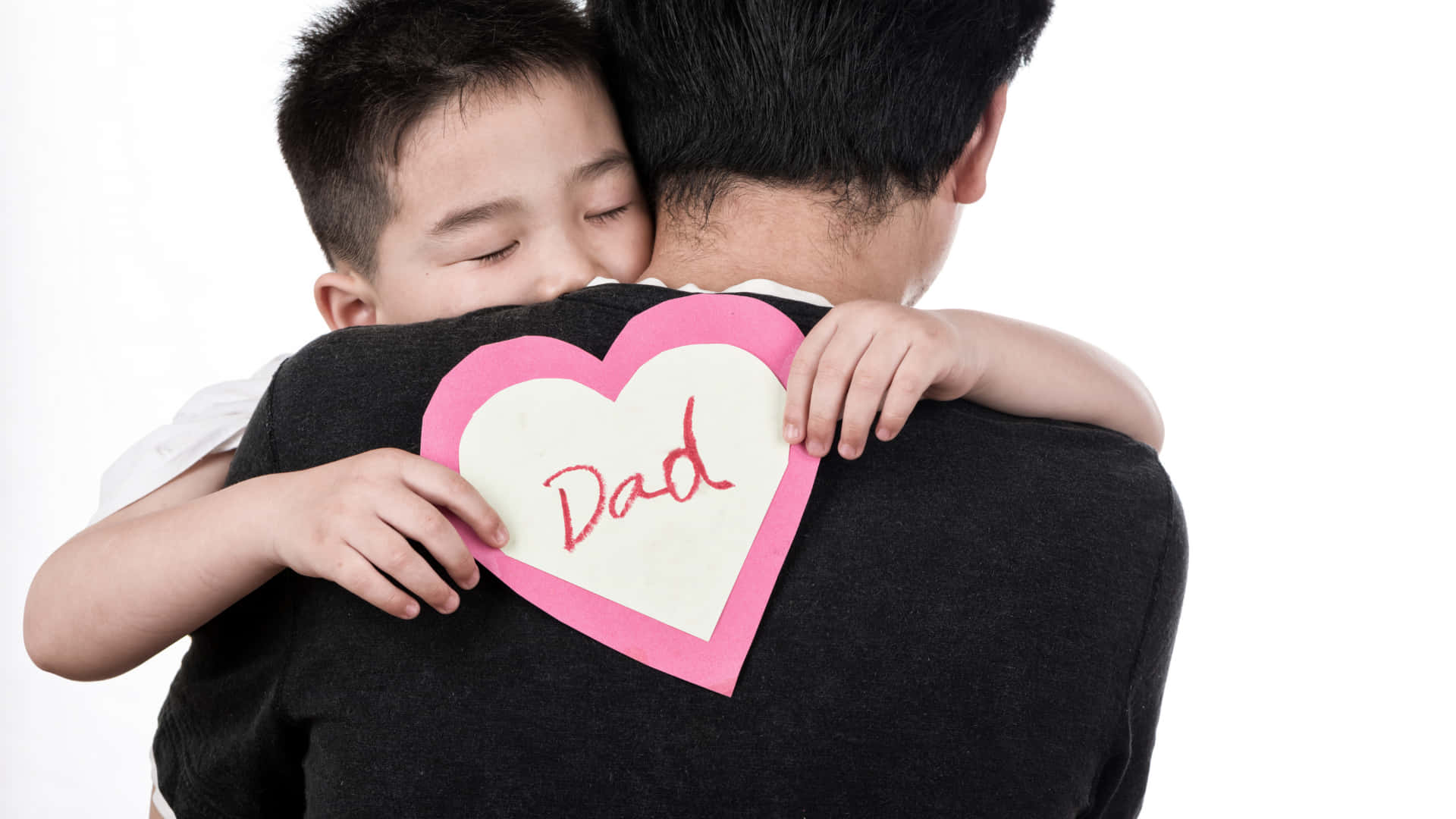 daddy 桌布 壁紙 — 一個小男孩從後面擁抱他的父親，同時拿著一個粉紅色的紙心，上面用紅色草書寫著「爸爸」。