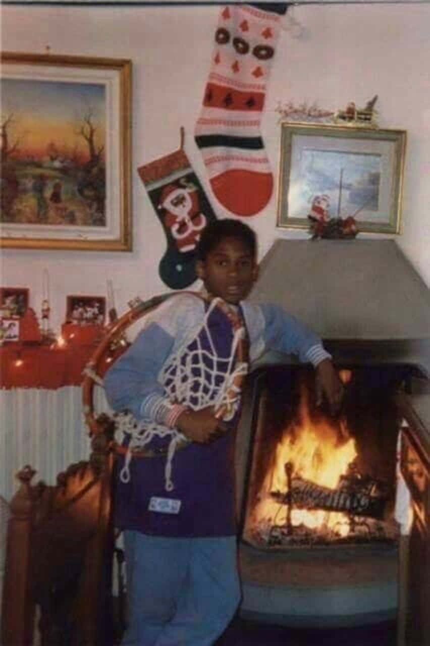 NBA聖誕節 桌布 壁紙 — 一個小男孩站在一個熊熊燃燒的壁爐旁邊，房間裝飾溫馨，掛滿聖誕襪和裱框藝術品。