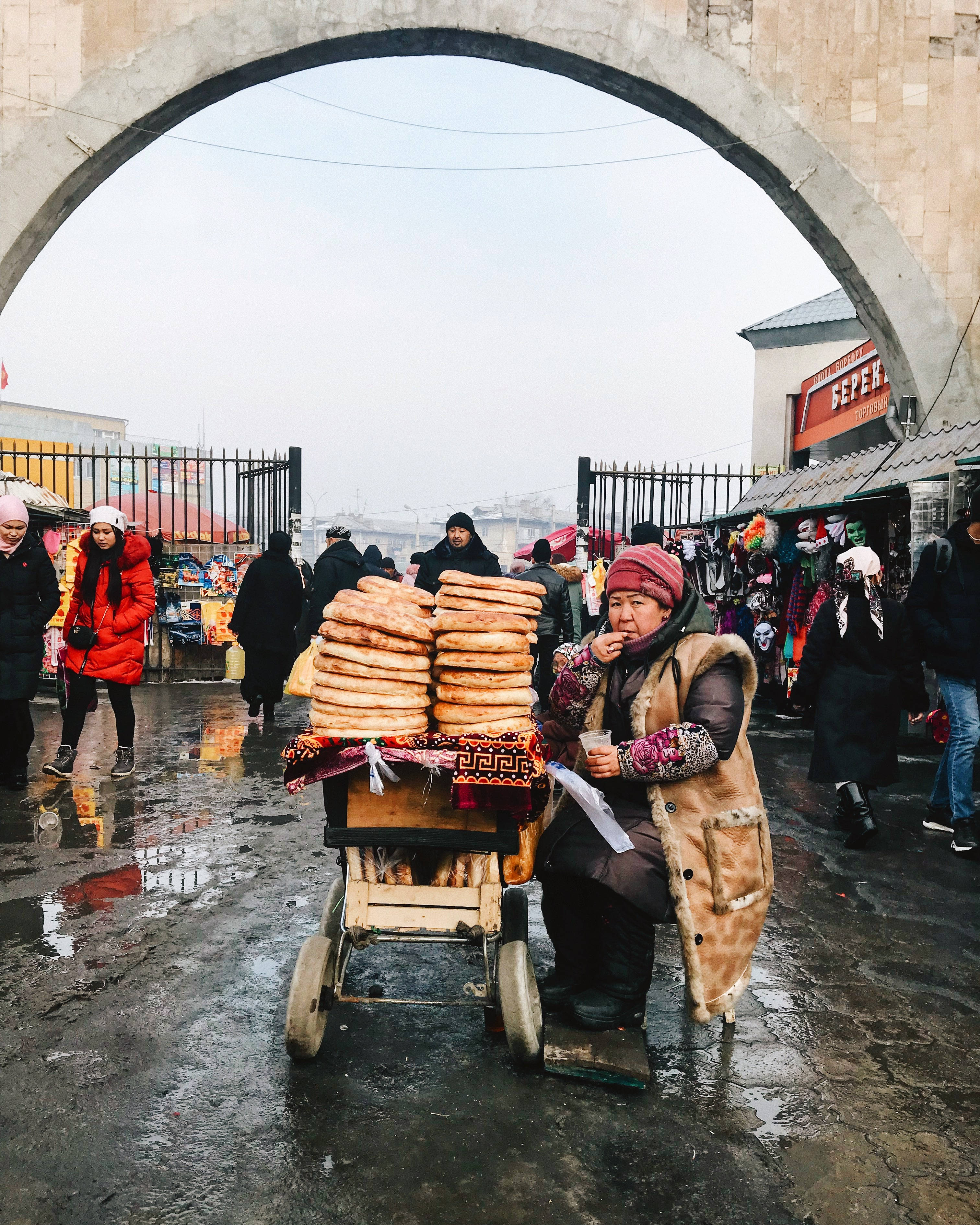 Kyrgyzstan 桌布 壁紙 — 一個小販坐在石拱門下，在潮濕的市場街道上，推車上賣著疊起的扁麵包。