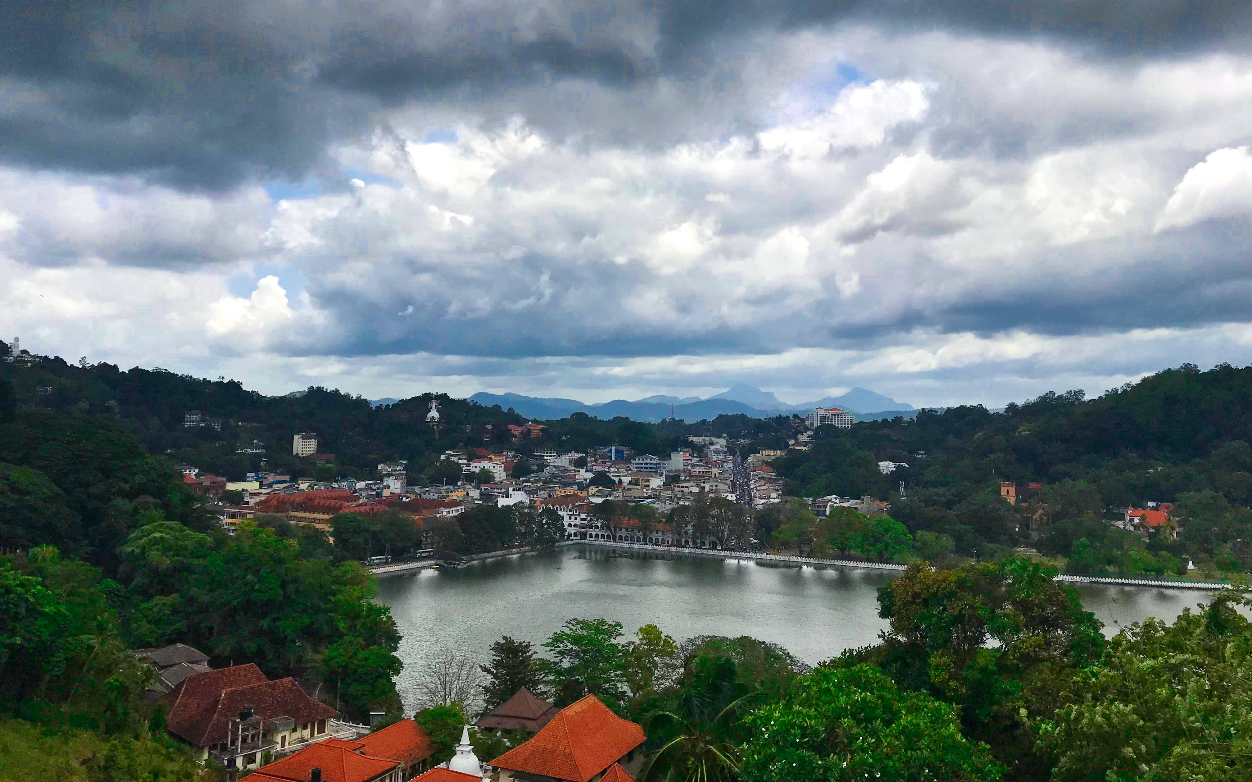 Sri Lanka 桌布 壁紙 — 一個小鎮坐落在平靜的湖泊周圍，天空戲劇性地多雲，背景是鬱鬱蔥蔥的綠色山丘。
