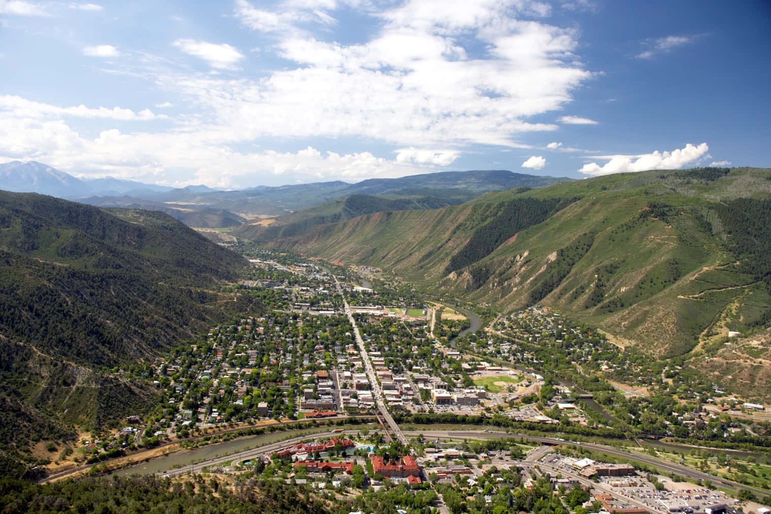 Colorado Mountain 桌布 壁紙 — 一個小鎮的鳥瞰圖，坐落於綠色山谷中，周圍環繞著起伏的丘陵和遠處白雪皚皚的山脈，天空部分多雲，藍天白雲。