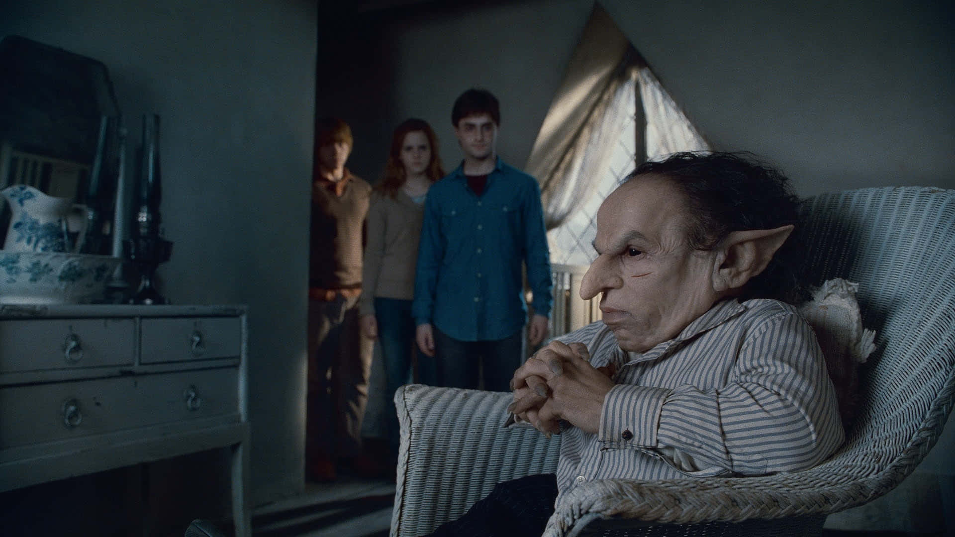 Warwick Davis 桌布 壁紙 — 一個尖耳朵的老妖精坐在陰暗房間的藤椅上，後面門口站著三個年輕人。