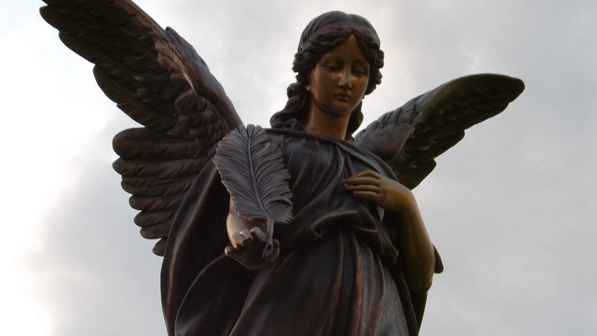 angels 桌布 壁紙 — 一個展開翅膀的青銅天使雕像，在陰雲密佈的天空下拿著一根羽毛。