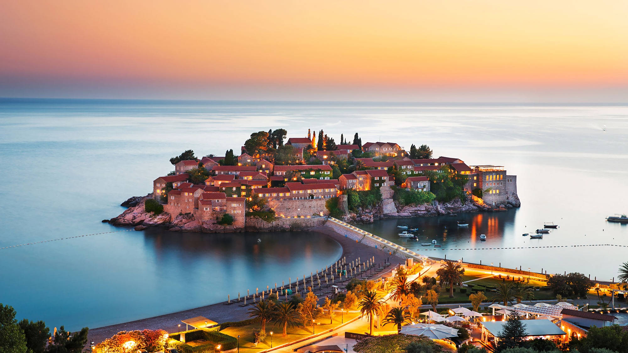 Montenegro 桌布 壁紙 — 一個岩石島上的沿海小鎮在日落時溫暖地發光，平靜的藍色海水和一條長長的燈光碼頭延伸向海岸。