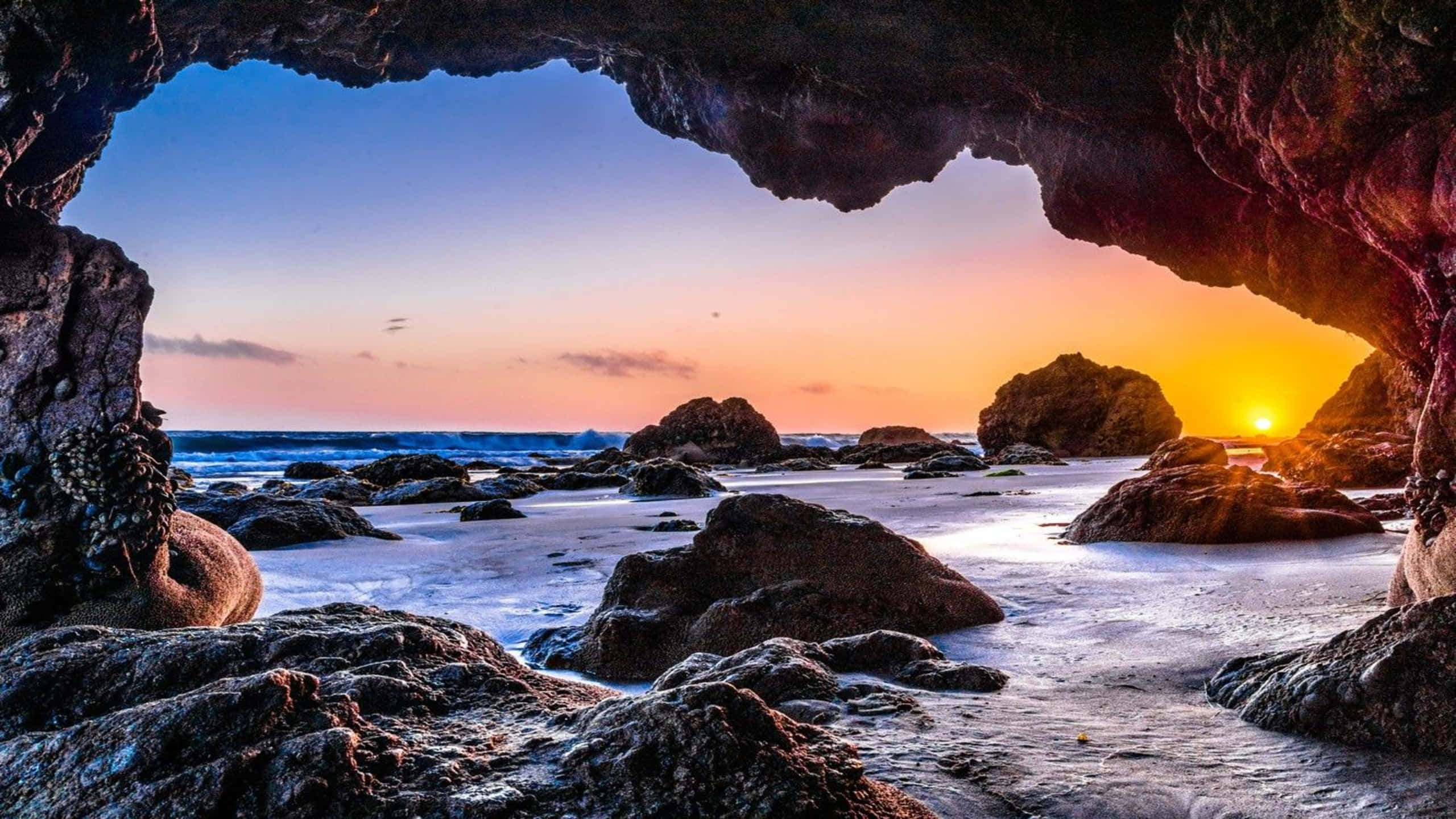 Rocks 桌布 壁紙 — 一個岩石海蝕洞框住了一個充滿活力的日落，橙色太陽在海洋波浪和散落的巨石上發光。