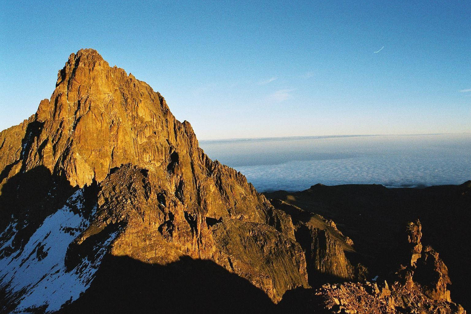 Kenya 桌布 壁紙 — 一個崎嶇的山峰沐浴在金黃色的日出光芒中，聳立在雲海之上，陰影籠罩的山坡上還掛著積雪。