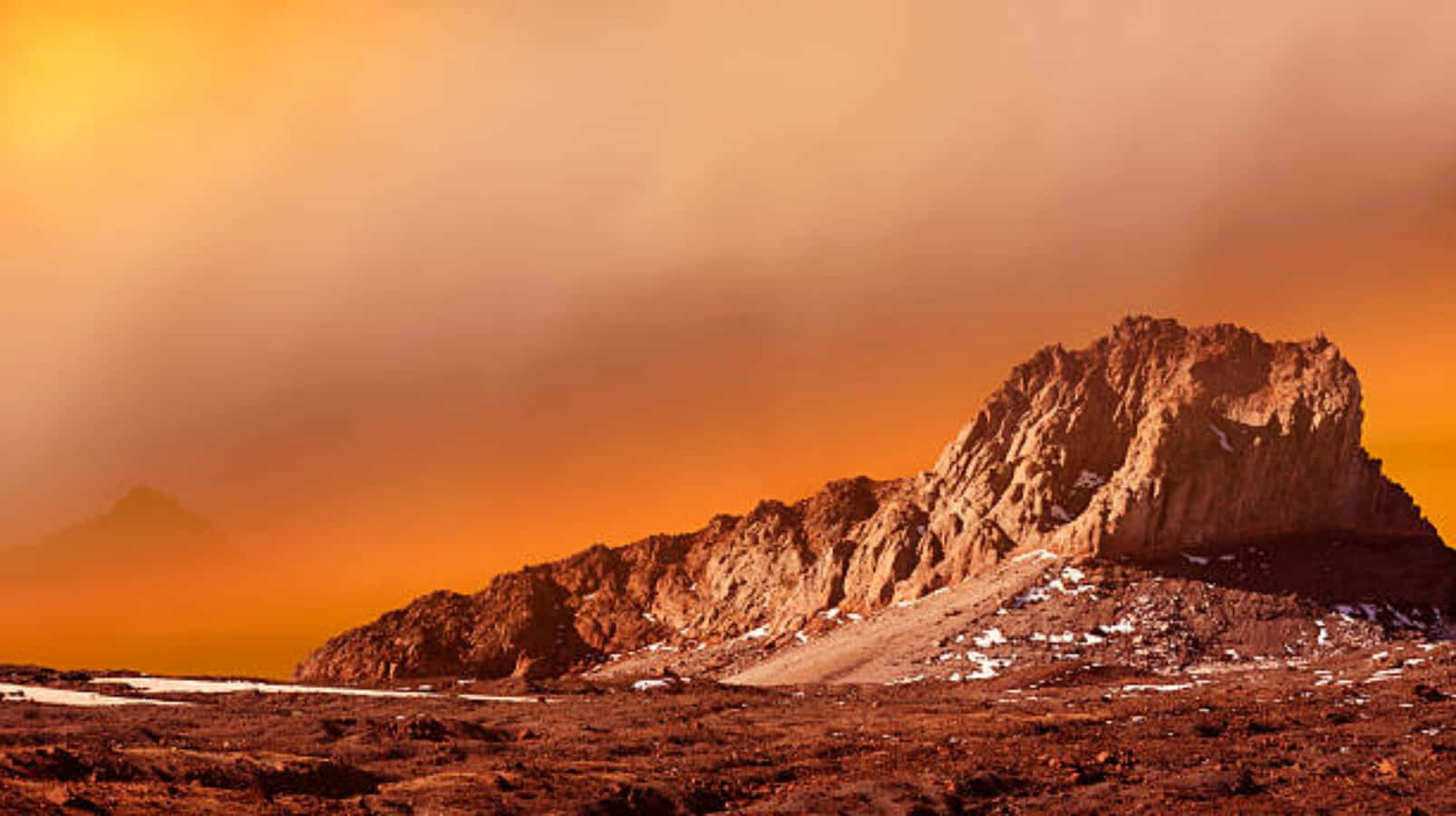 Mars Surface 桌布 壁紙 — 一個崎嶇的岩石山峰，在朦朧的日落天空下，沐浴在溫暖的橙色光芒中，散落著雪斑。