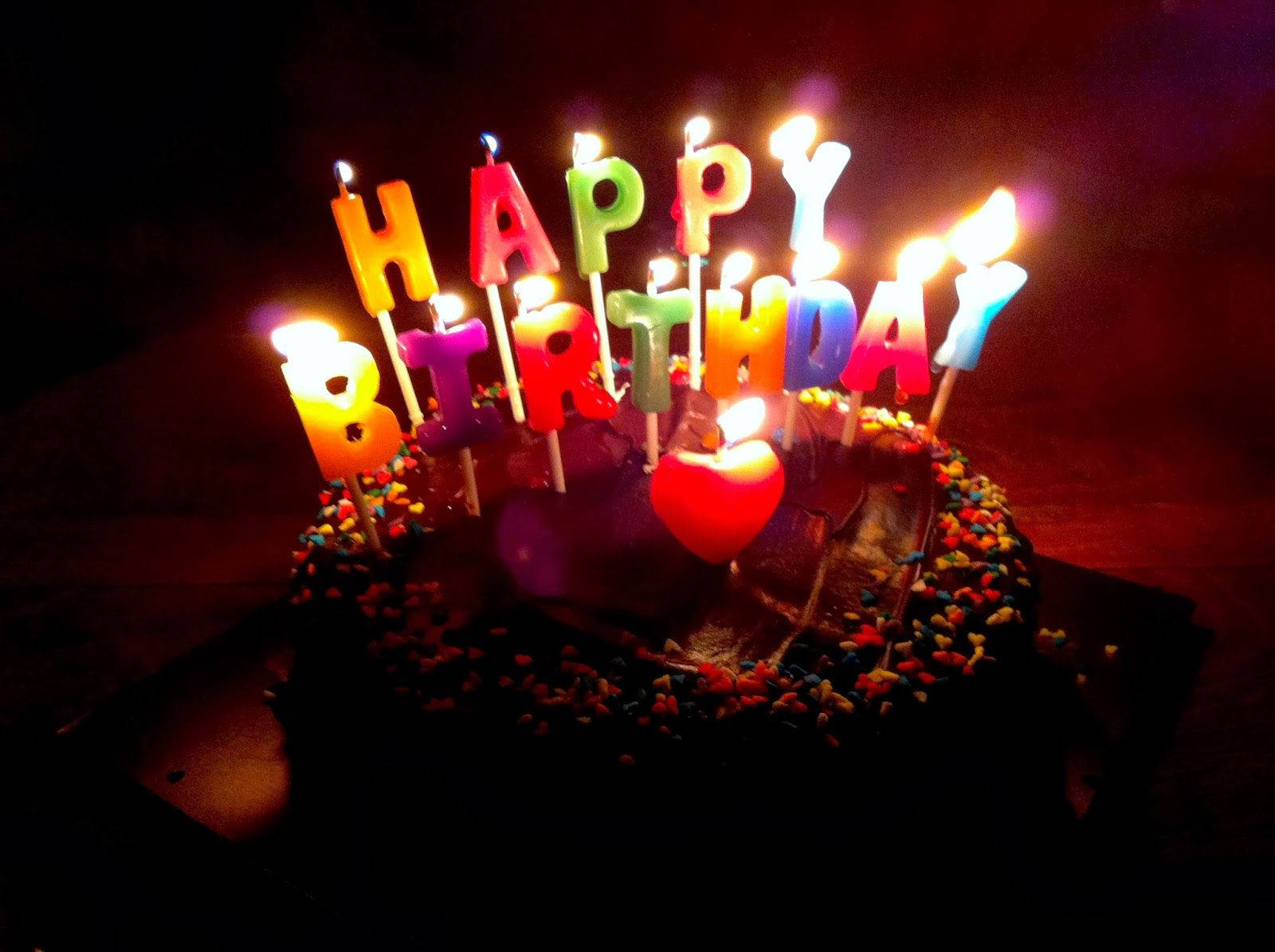 Birthday 桌布 壁紙 — 一個巧克力生日蛋糕，上面有色彩繽紛的點燃蠟燭拼出 happy birthday，還有一顆心形蠟燭在黑暗中發光。