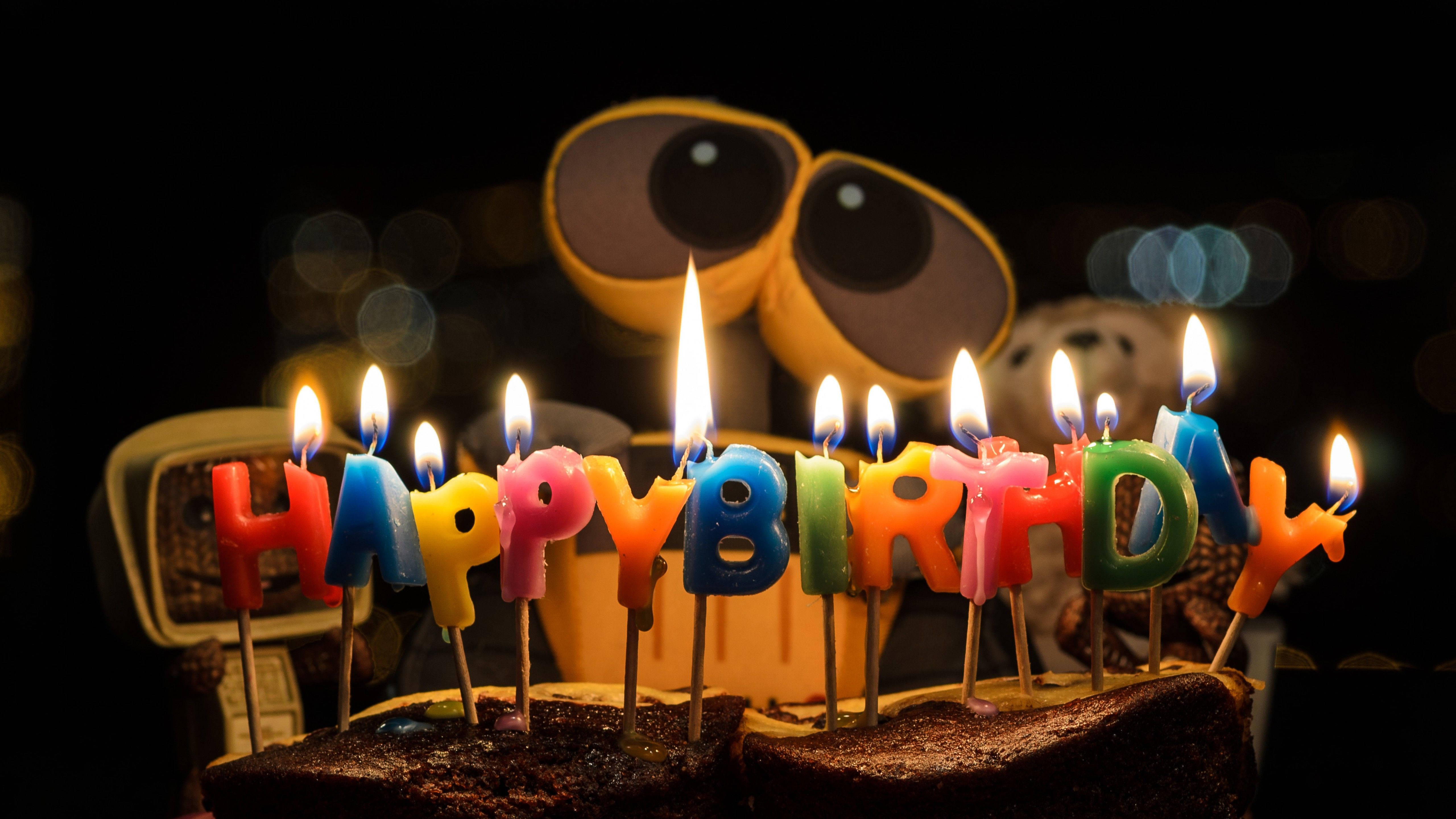 生日快樂 桌布 壁紙 — 一個巧克力生日蛋糕，上面有色彩繽紛的點燃蠟燭拼出 happy birthday，放在一個大眼睛機器人玩具前面，背景是暗色散景。