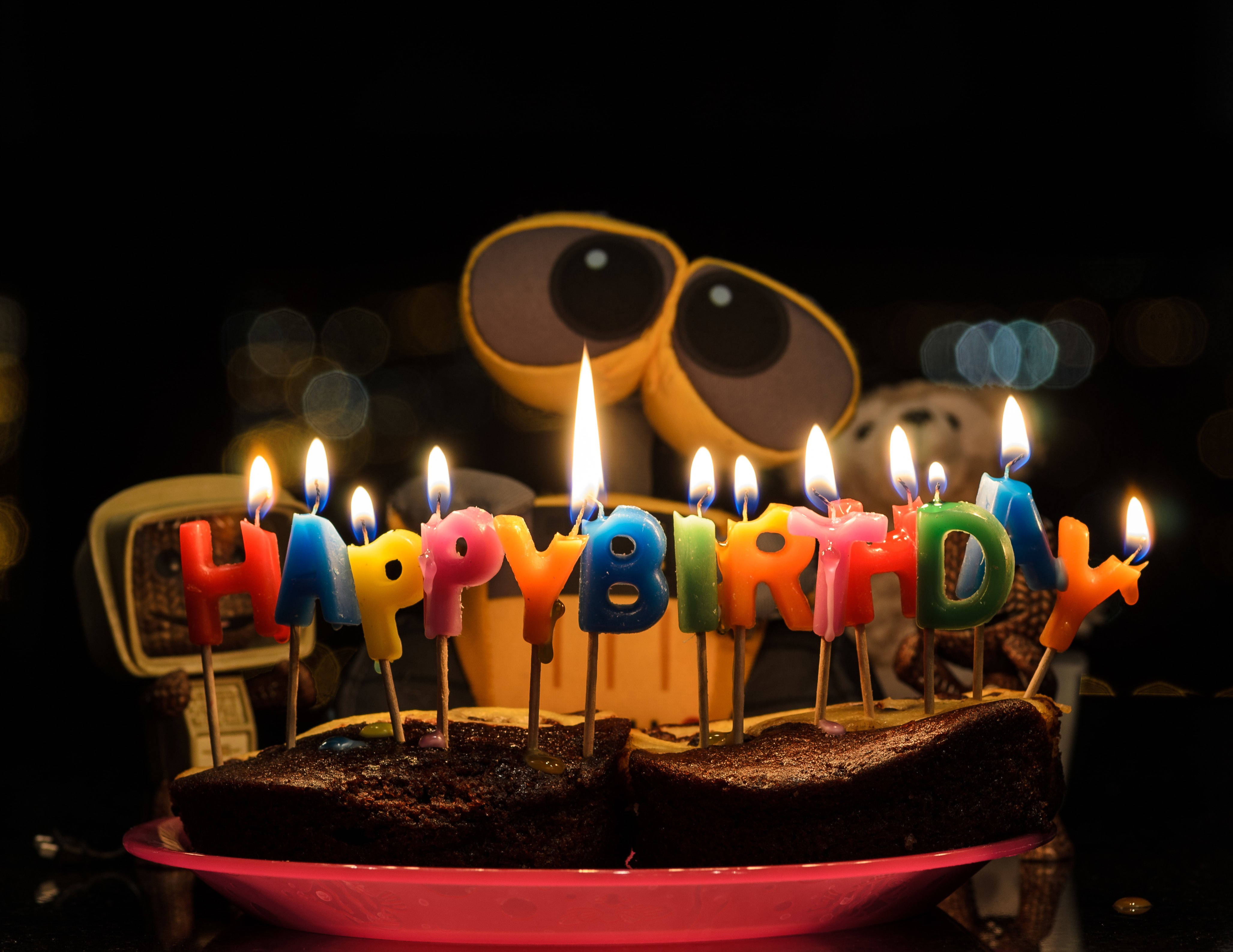 E 桌布 壁紙 — 一個巧克力生日蛋糕，前面有色彩繽紛的「生日快樂」蠟燭點燃，背景黑暗，前面有一個大型的Wall-E絨毛玩具。