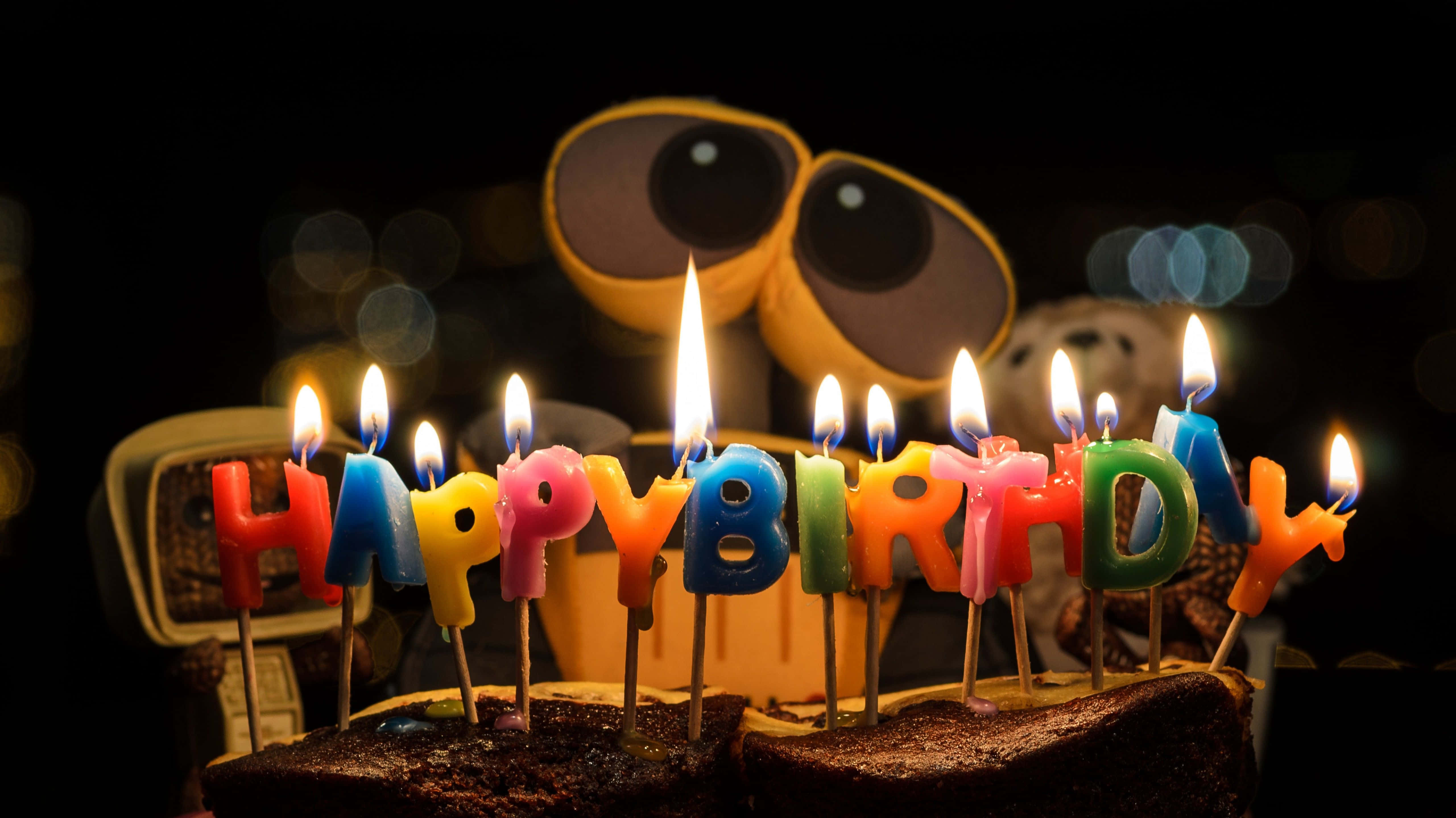 Birthday 桌布 壁紙 — 一個巧克力生日蛋糕，帶有色彩繽紛的「生日快樂」蠟燭，在黑暗背景中閃耀，後面有一個大眼睛機器人玩具。