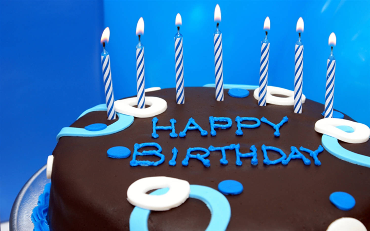 Cake 桌布 壁紙 — 一個巧克力生日蛋糕，帶有藍白條紋蠟燭和 'HAPPY BIRTHDAY' 糖霜，背景是鮮豔的藍色。