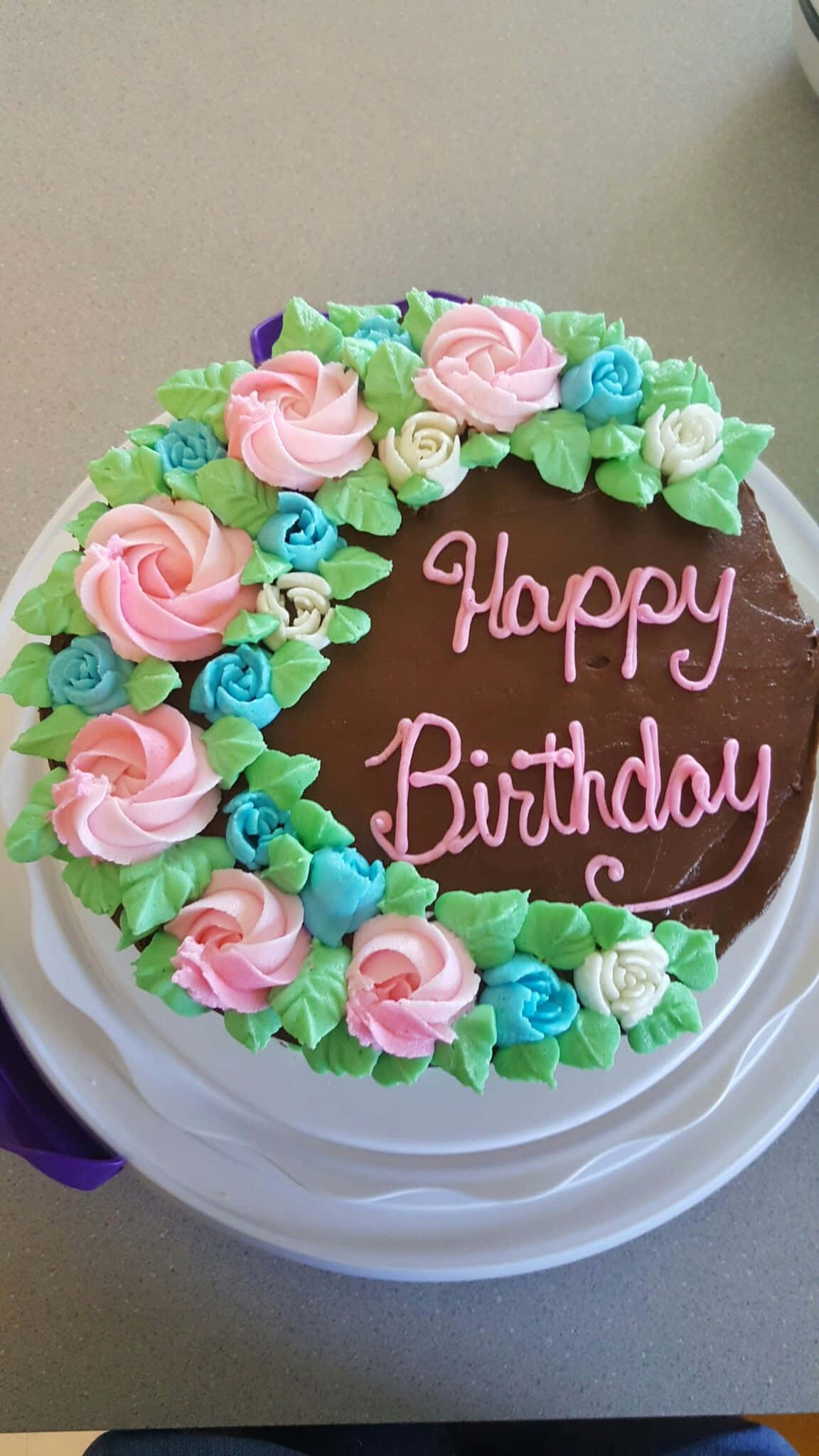 Happy Birthday Cake 桌布 壁紙 — 一個巧克力生日蛋糕，裝飾有粉紅色、藍色和白色的糖霜玫瑰和綠色葉子，並有粉紅色草書Happy Birthday文字。