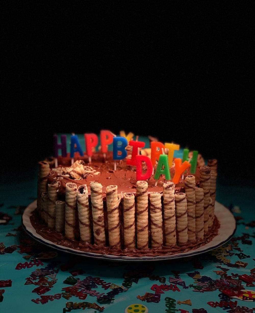 Birthday Cake 桌布 壁紙 — 一個巧克力生日蛋糕，飾以彩色字母蠟燭和威化卷，放在鋪滿五彩紙屑的桌子上，在戲劇性的燈光下。