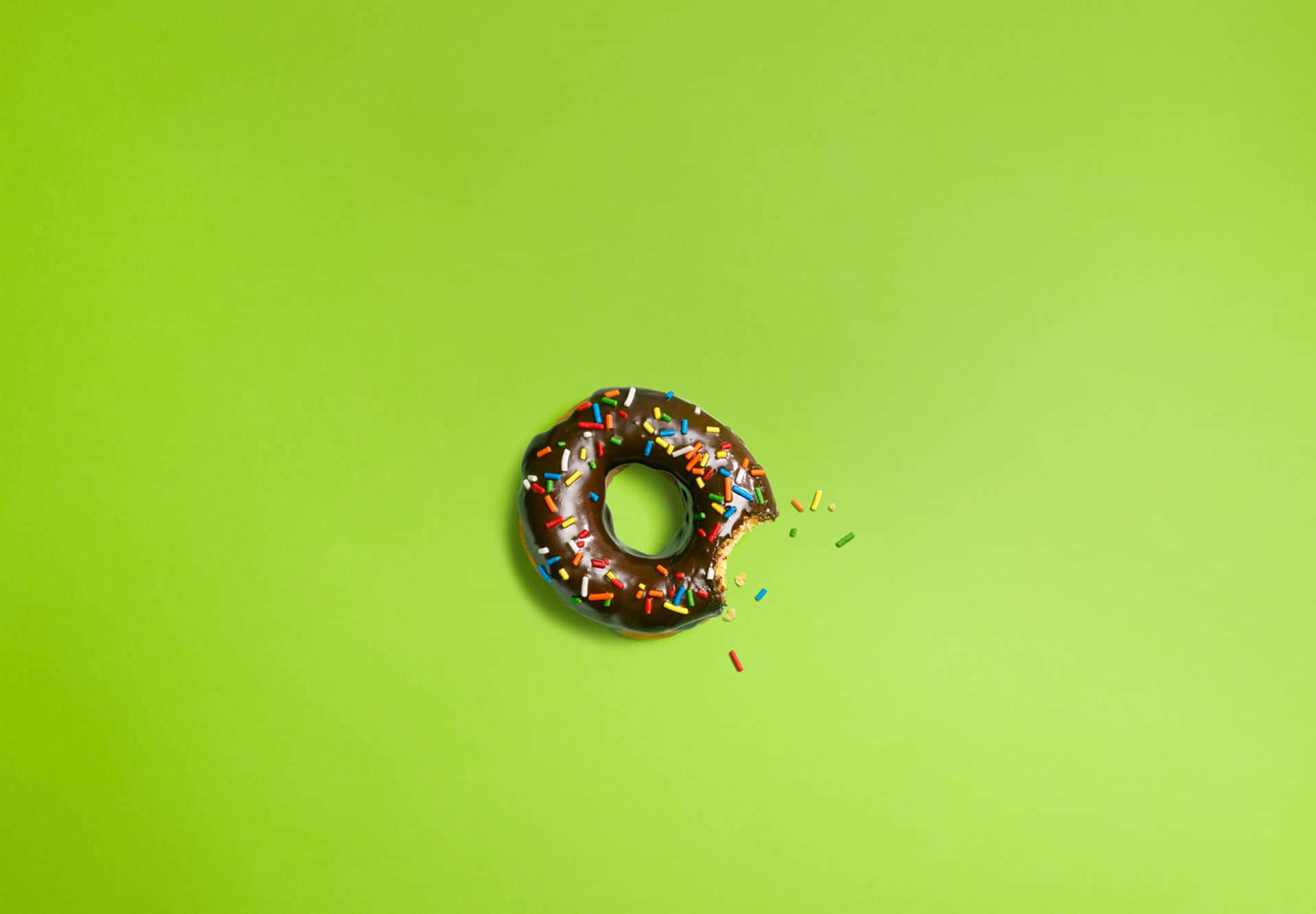 donuts 桌布 壁紙 — 一個巧克力糖霜甜甜圈，上面有彩色糖粒，放在鮮綠色背景上，被咬了一口。