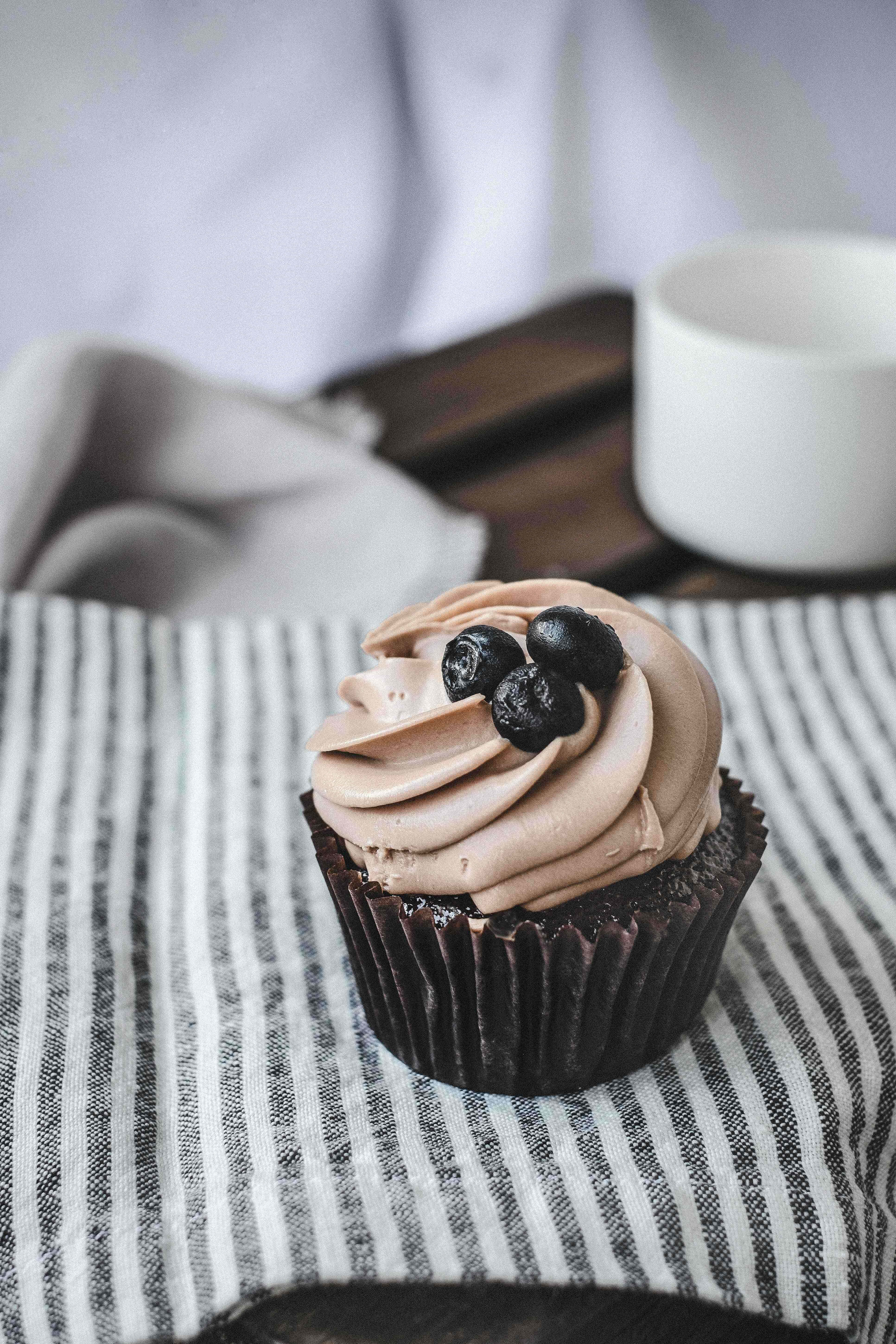 Cupcake 桌布 壁紙 — 一個巧克力紙杯蛋糕，上面有淺棕色糖霜漩渦和新鮮藍莓，放在黑白條紋布上。