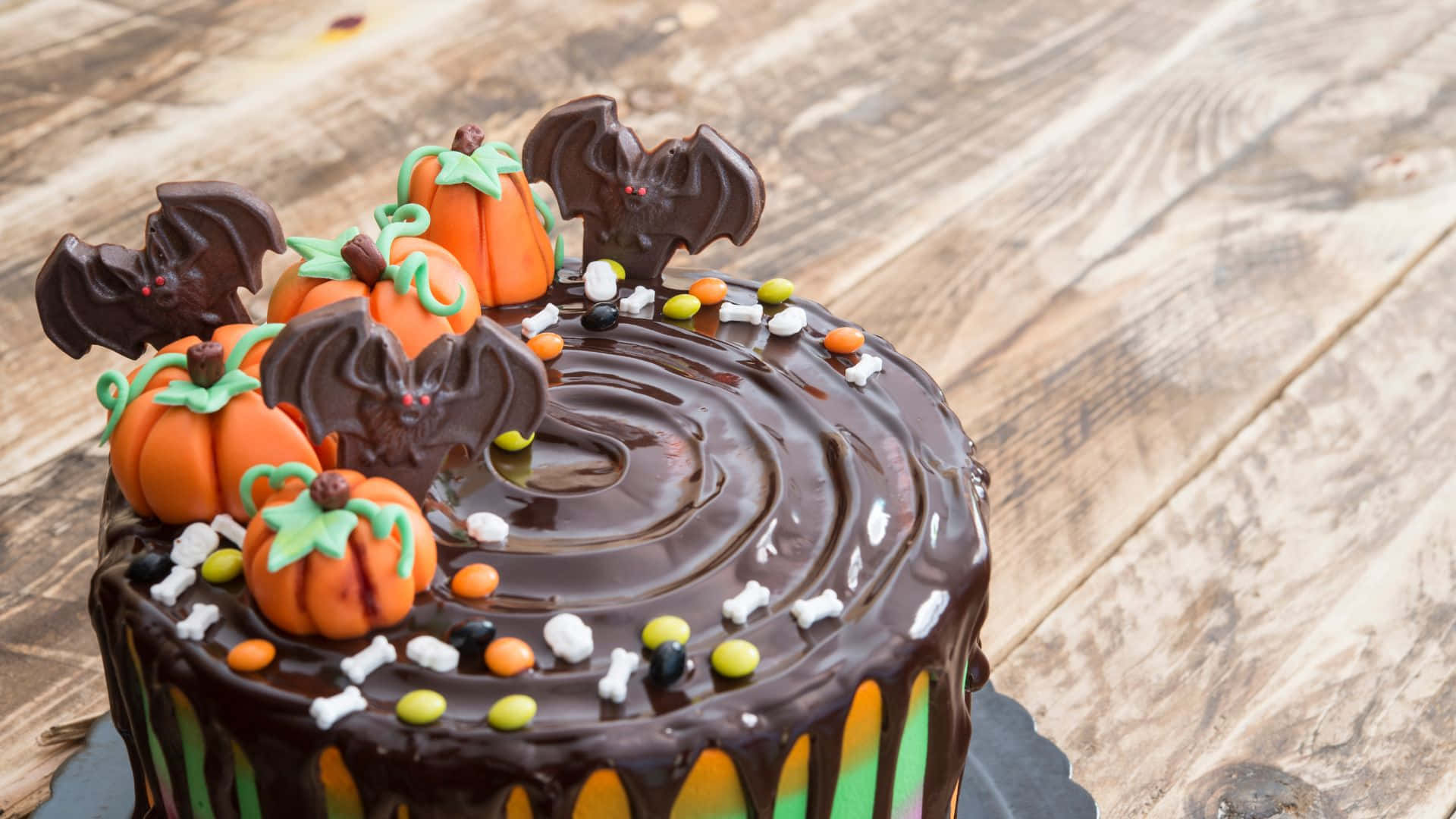 Halloween Cake 桌布 壁紙 — 一個巧克力萬聖節蛋糕，裝飾著橙色南瓜、巧克力蝙蝠和糖果骨頭，放在質樸的木桌上。