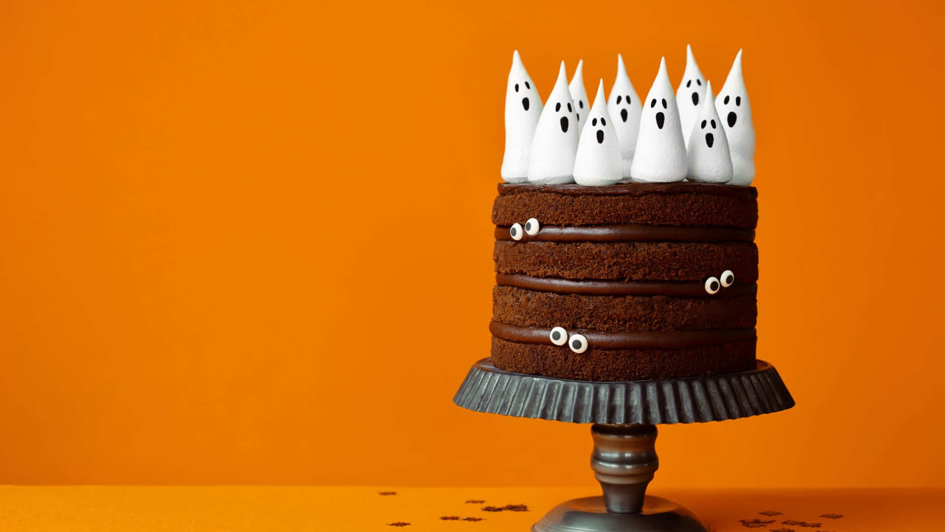 Halloween Cake 桌布 壁紙 — 一個巧克力蛋糕，飾以白色鬼魂蛋白霜和糖果眼睛，放在復古架上，背景是鮮豔的橙色。