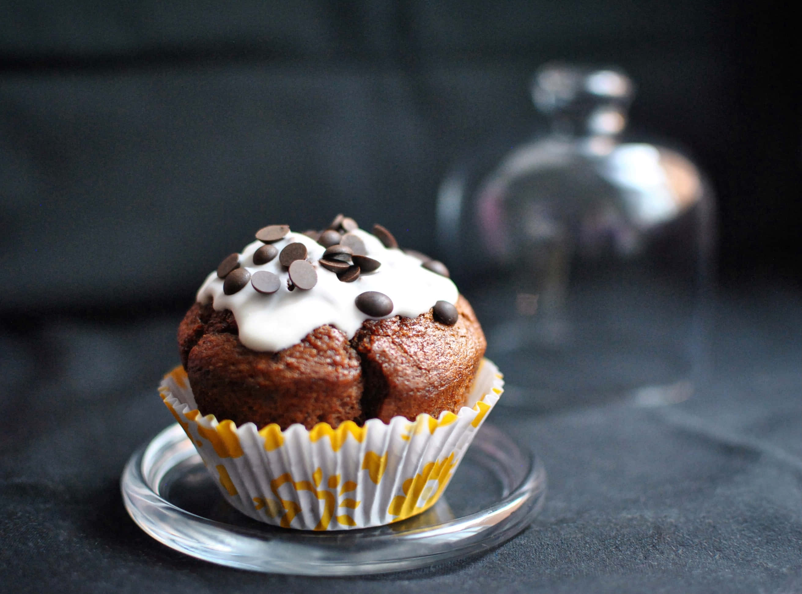 Cupcake 桌布 壁紙 — 一個巧克力鬆餅，上面有白色糖霜和巧克力豆，放在玻璃盤上，背景是深色有紋理的。