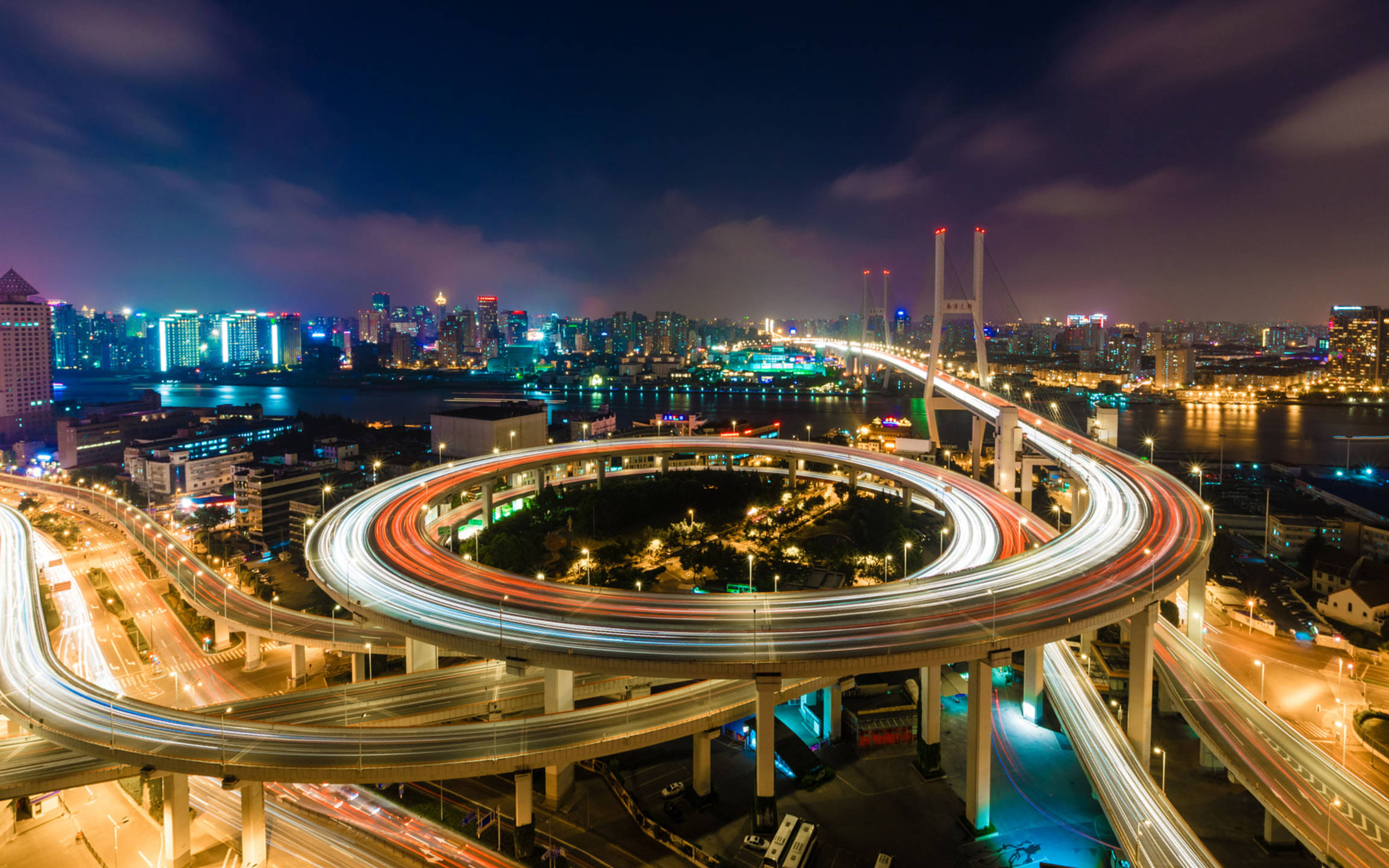 風景 桌布 壁紙 — 一個巨大照明高速公路交匯處的迷人夜景，車流的光跡映襯著閃爍的城市天際線。