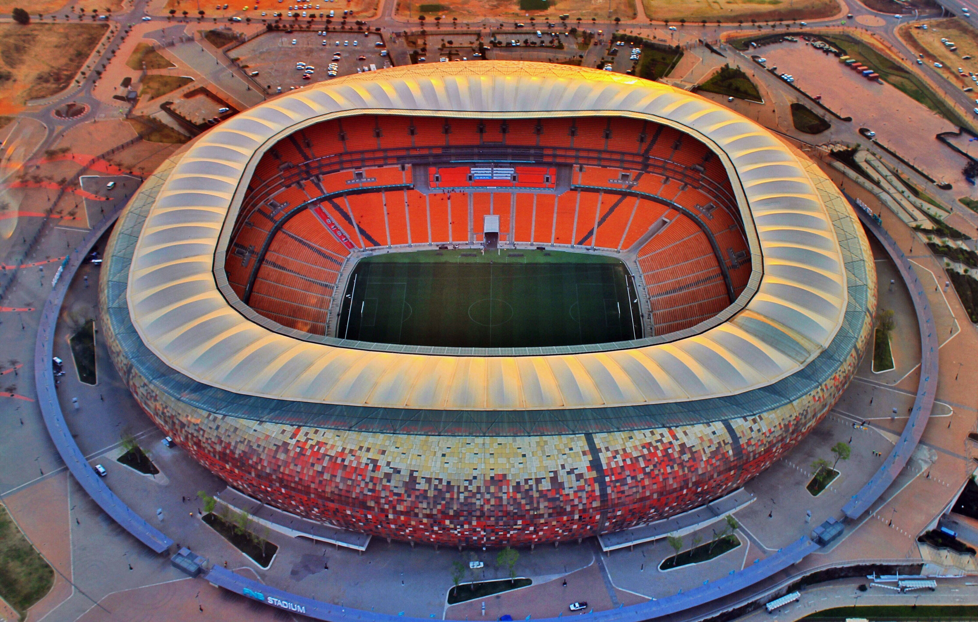 Stadium 桌布 壁紙 — 一個巨大現代體育場的鳥瞰圖，其鮮豔的紅色和金色外牆在金色時光下閃耀。