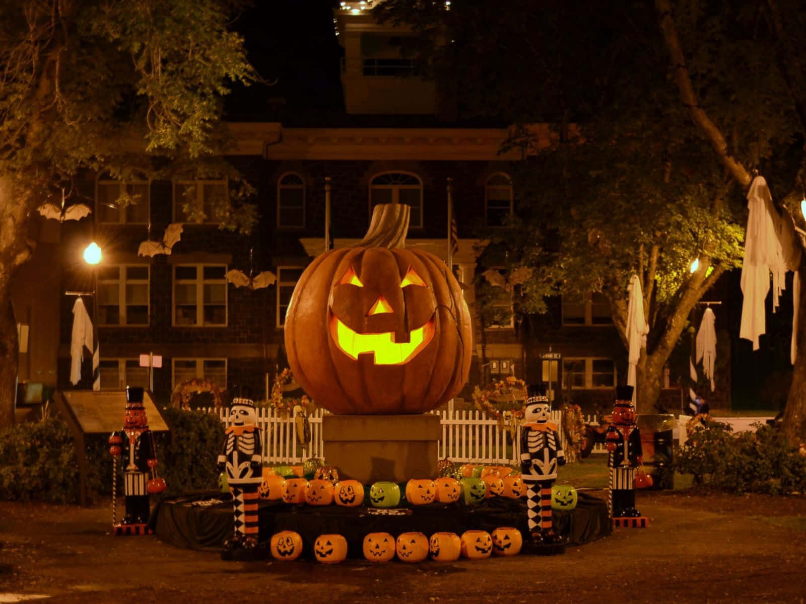 Halloweentown 桌布 壁紙 — 一個巨大發光的南瓜燈，咧嘴大笑，坐在基座上，周圍環繞著較小的南瓜和胡桃夾子骷髏，在夜燈下。
