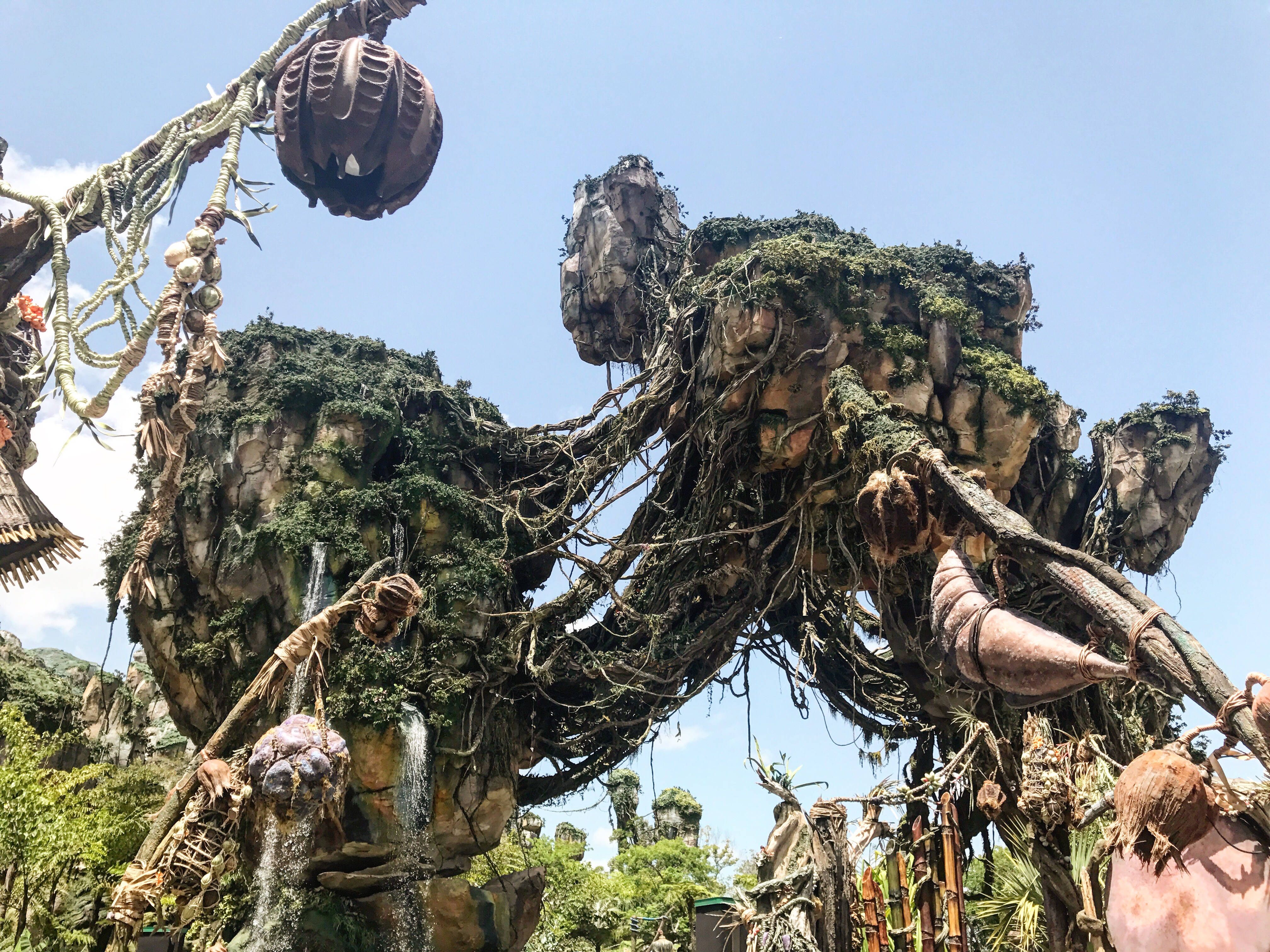 潘朵拉 桌布 壁紙 — 一個巨大的、長滿苔蘚的樹狀結構，帶有粗糙的根部和懸掛的豆莢，在晴朗的藍天下高聳入雲。