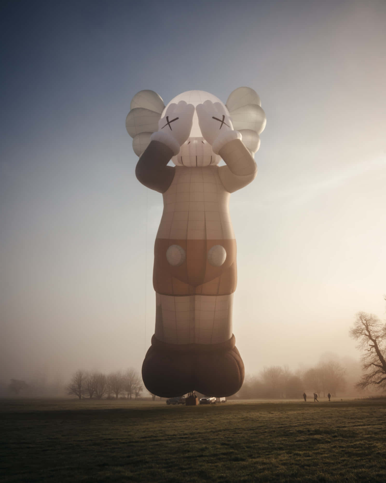 KAWS 桌布 壁紙 — 一個巨大的KAWS氣球雕塑在日出時分矗立於霧氣瀰漫的田野中，遠處有人們在觀望。