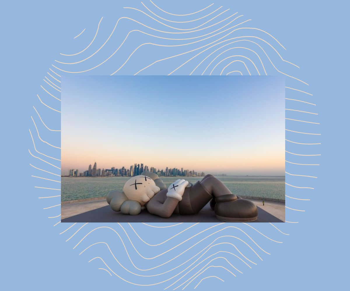 Kaws Holiday 桌布 壁紙 — 一個巨大的KAWS雕塑在海濱長廊上躺臥，夕陽下城市天際線閃耀著光芒。