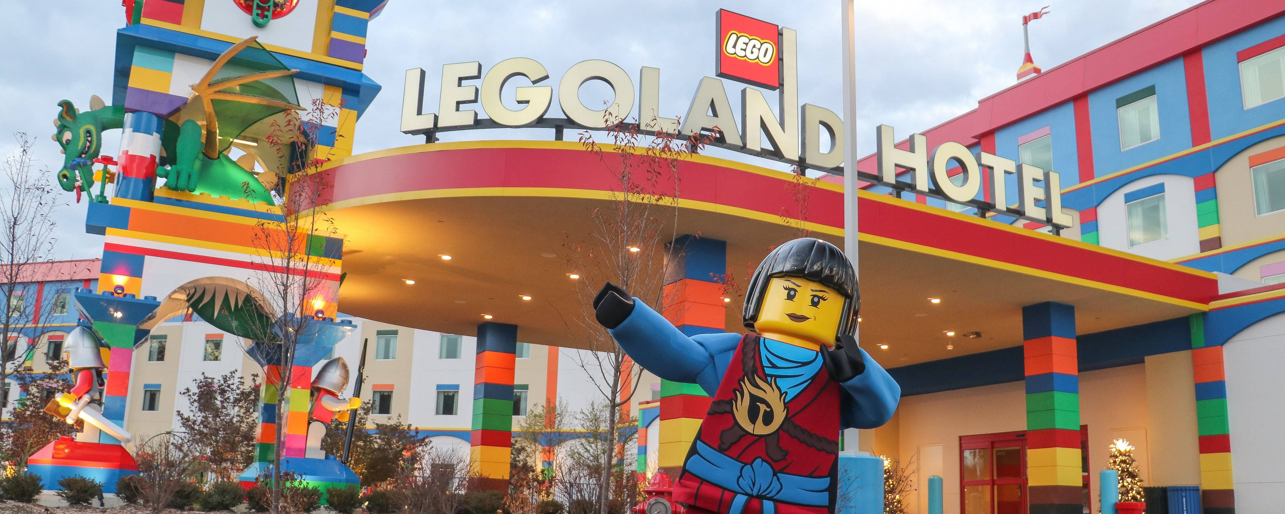飯店 桌布 壁紙 — 一個巨大的LEGO人偶在色彩繽紛的LEGO Land Hotel前愉快地揮手，酒店有彩虹磚柱和龍雕塑。