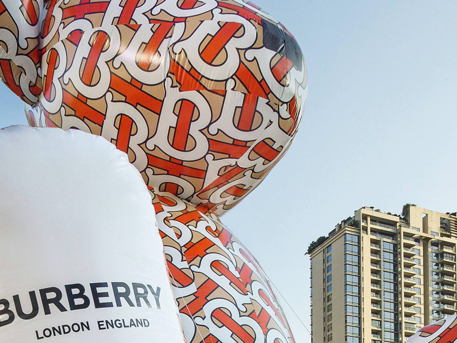 Burberry 桌布 壁紙 — 一個巨大的充氣 Burberry 標誌雕塑聳立在現代城市天際線之上，背景是晴朗的藍天。